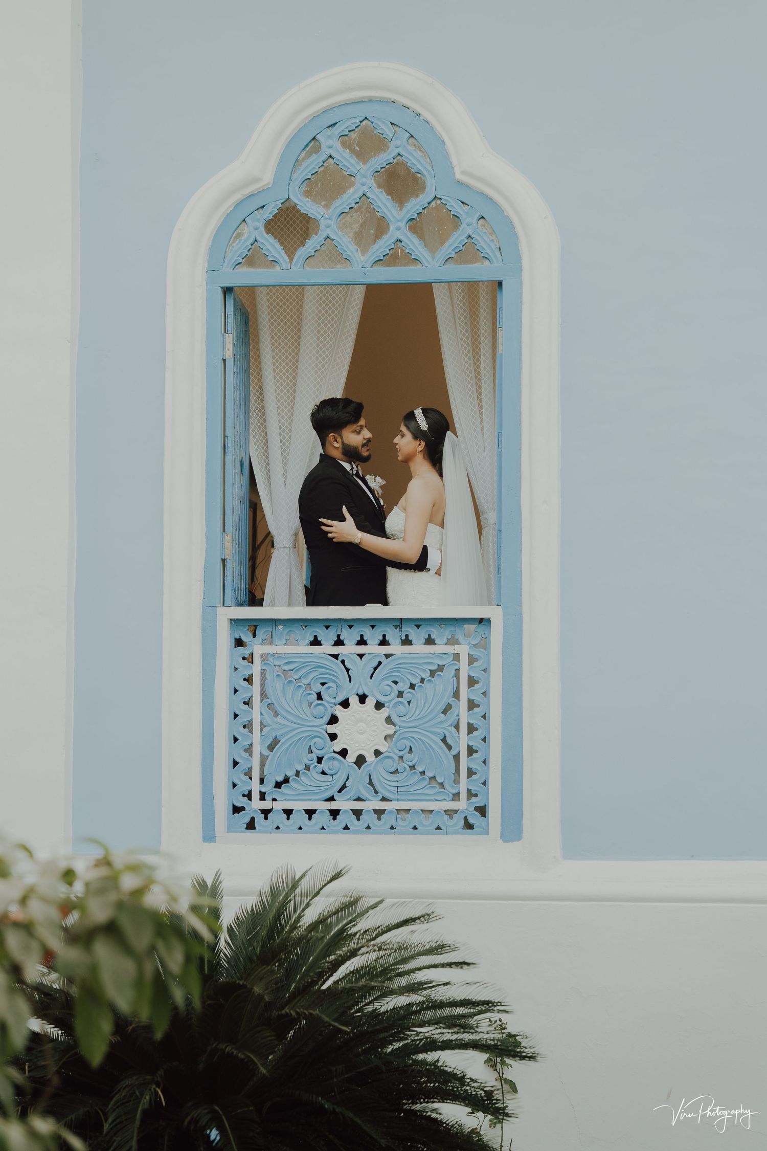 A romantic moment framed by an ornate blue and white Mediterranean-style window with decorative latticework and plants below.