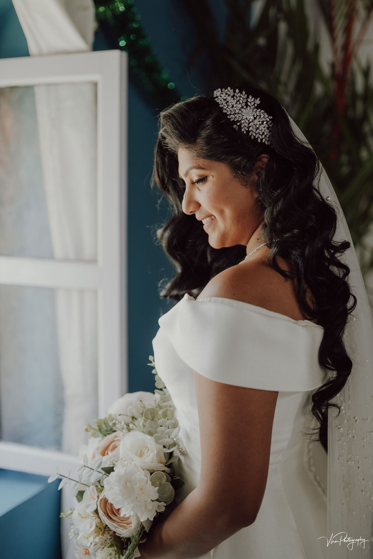 A bride in a classic off-shoulder white gown holds a cream and blush bouquet while wearing an elegant crystal hair accessory.