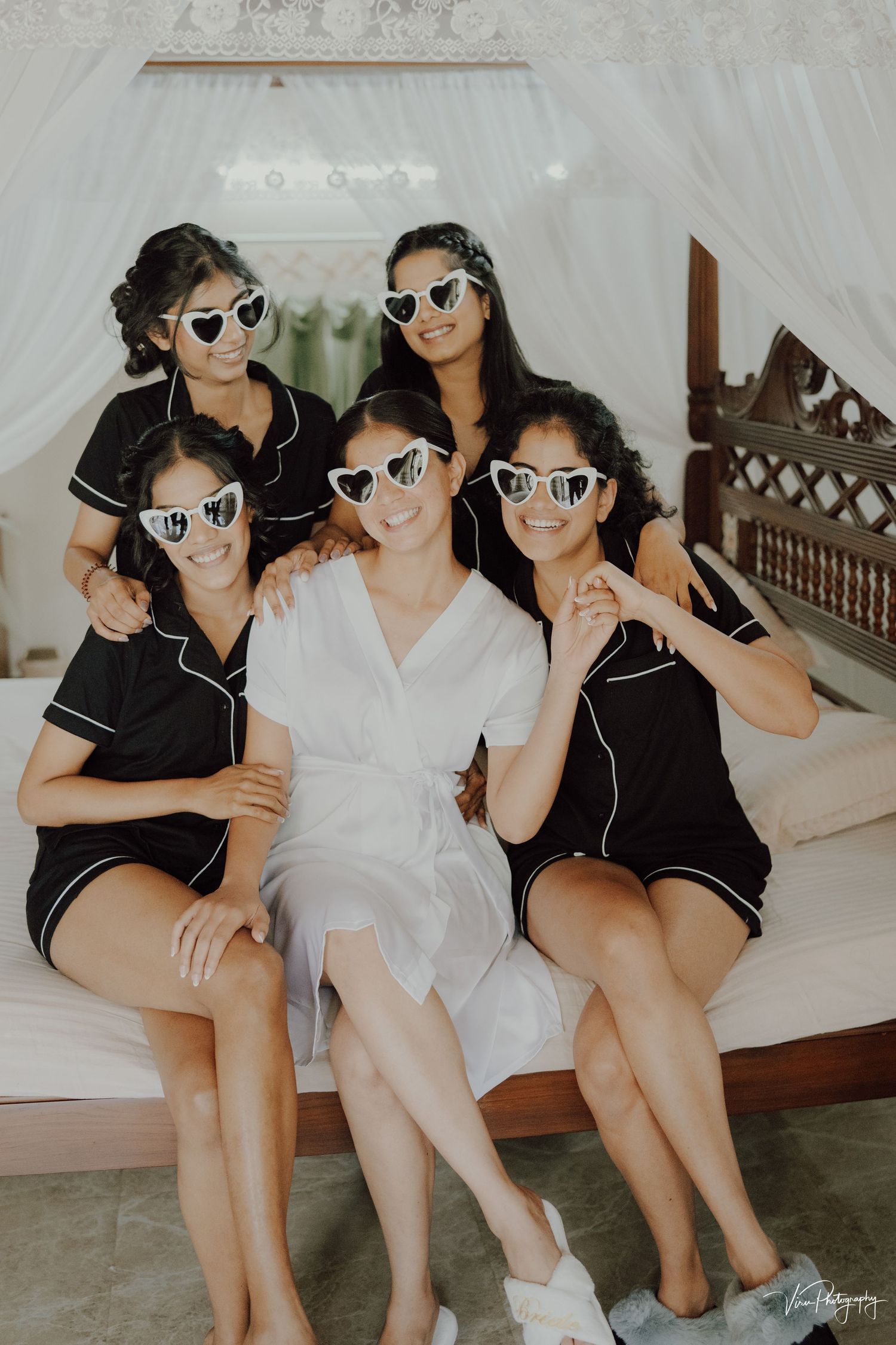 Five people sitting together on white bedding wearing matching mirrored sunglasses during pre-wedding preparations.