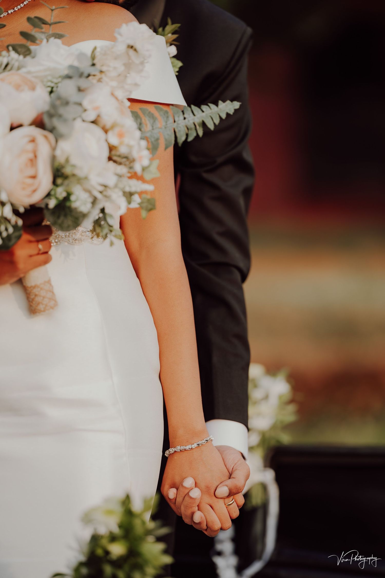 Close-up of newlywed couple holding hands with white bridal bouquet featuring roses and ferns in the background.