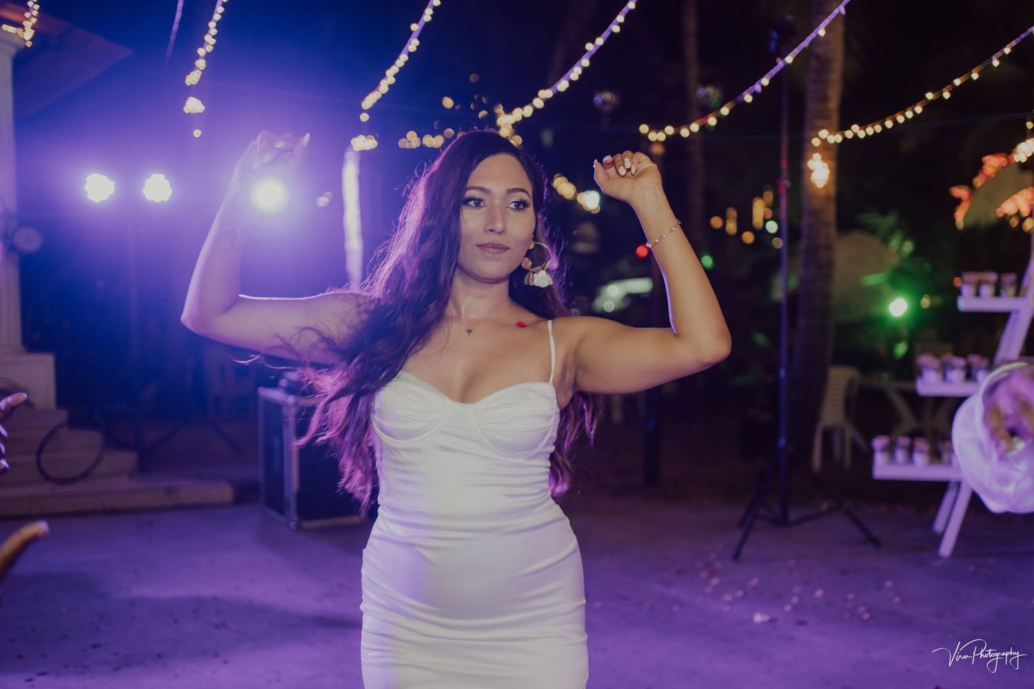 A person in a fitted white dress dances at an evening celebration with string lights and purple ambient lighting in the background.