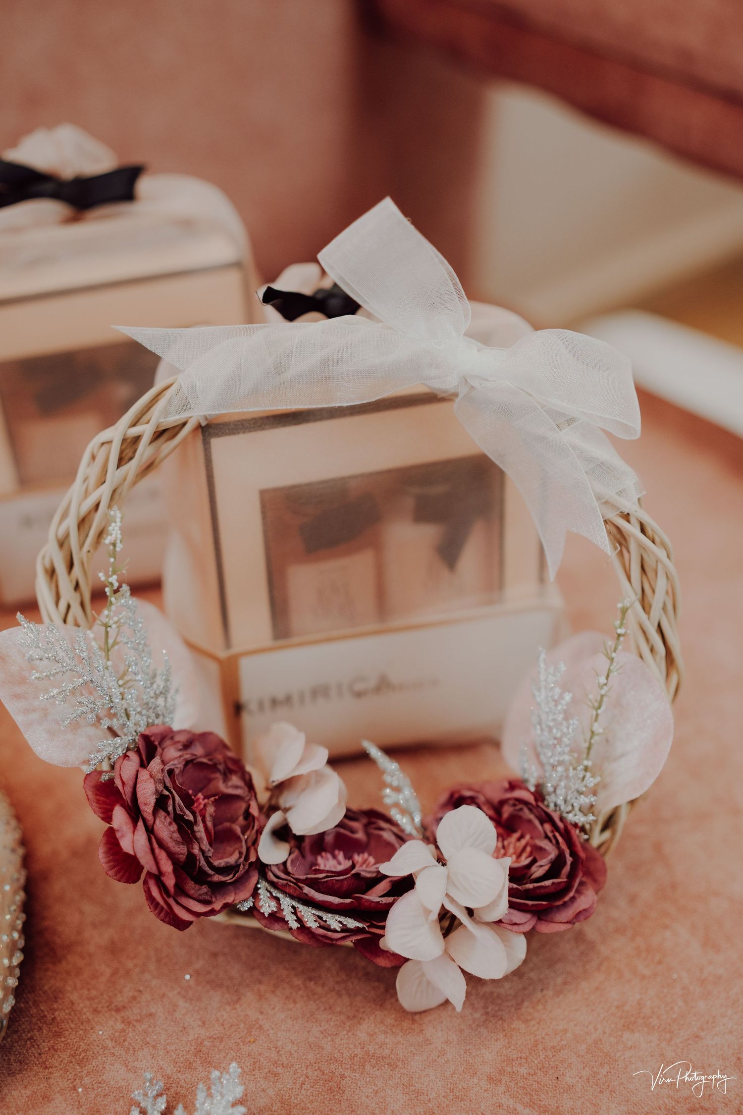 A decorative white wicker wreath adorned with burgundy roses, white flowers and a sheer bow sits near gift boxes.