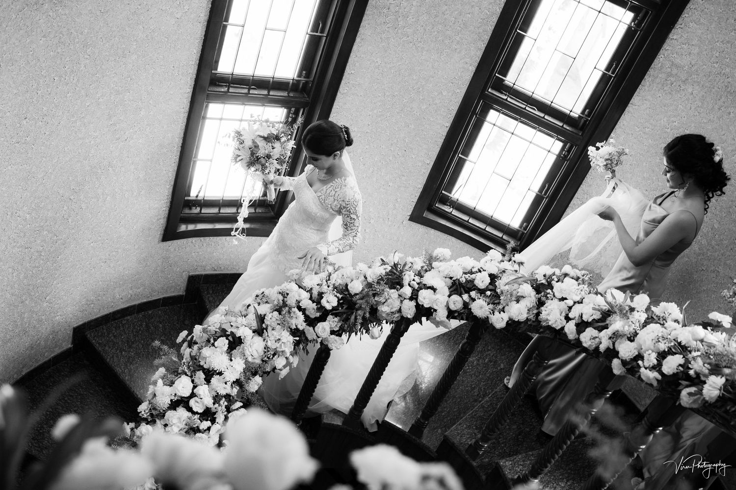 Black and white photograph showing a floral bridal arch while wedding preparations take place in a room with window light.