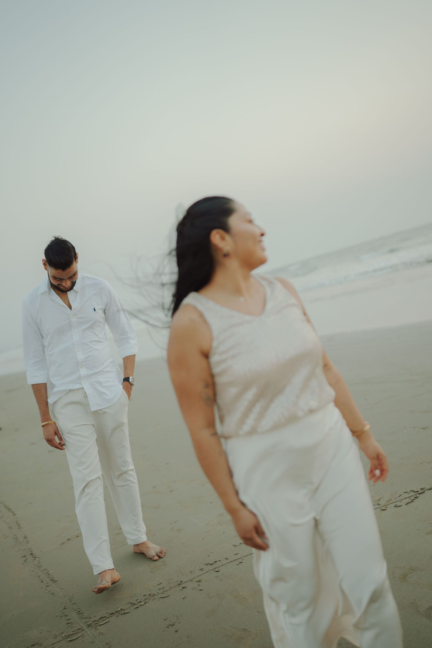 Couple walking barefoot on a misty beach at sunset, dressed in elegant white outfits.