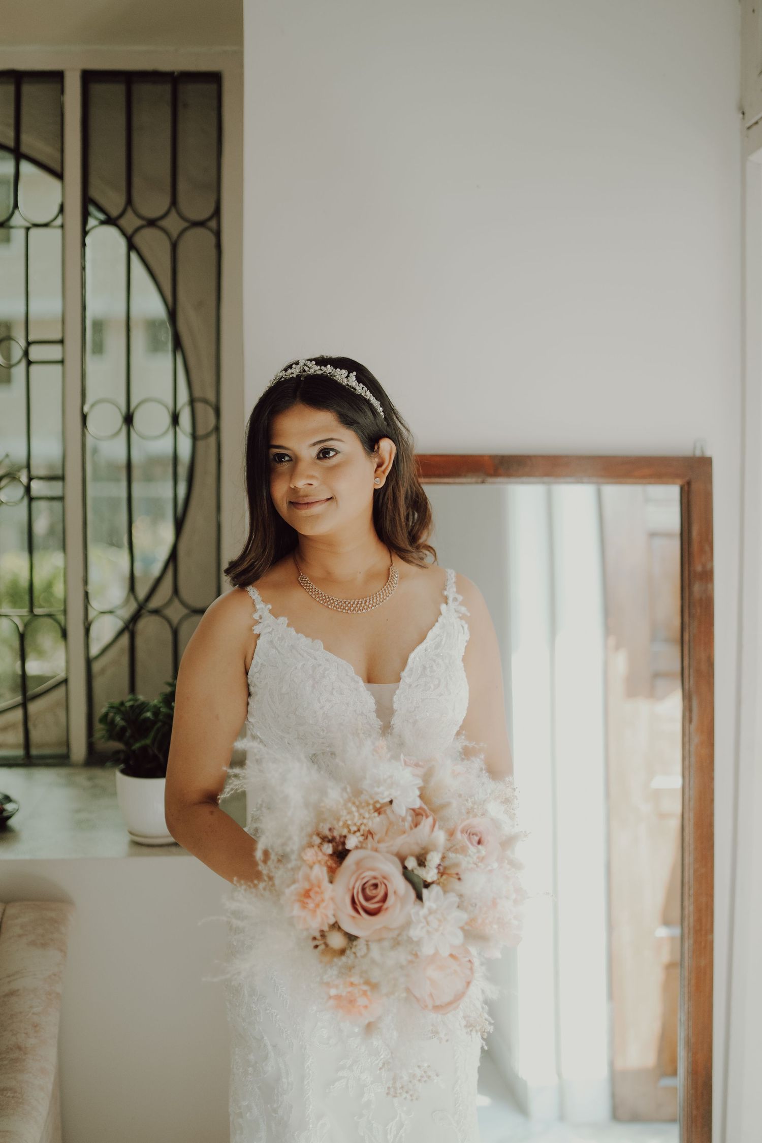 Radiant bride in fitted lace gown holding pink and white bouquet by window.