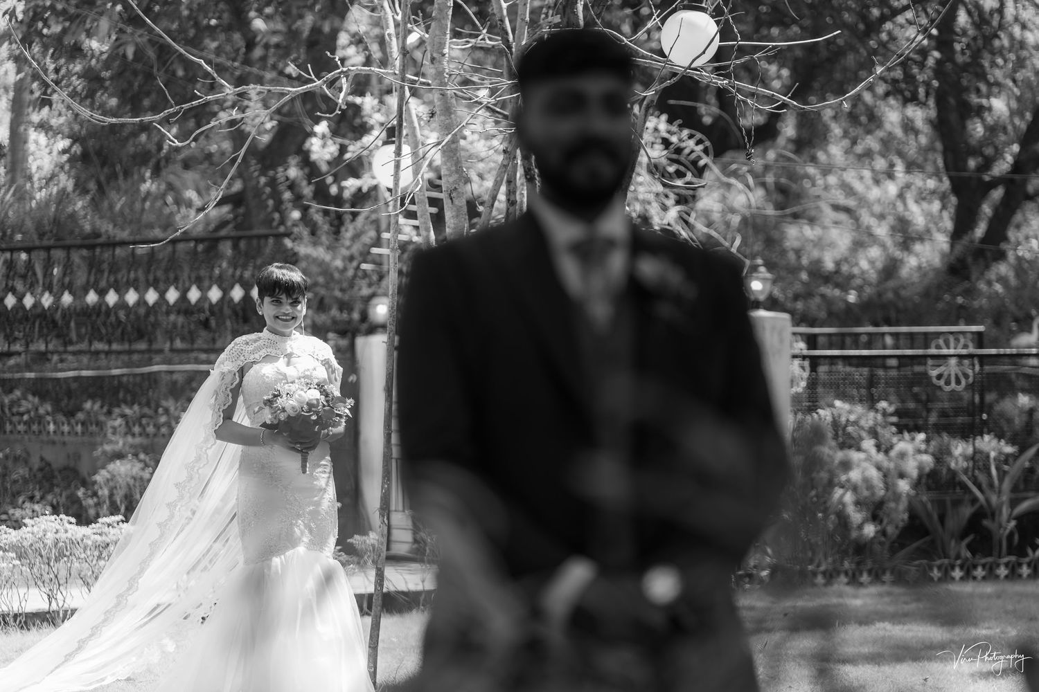 Artistic black and white wedding photograph showing a blurred groom in foreground and bride in background outdoors.