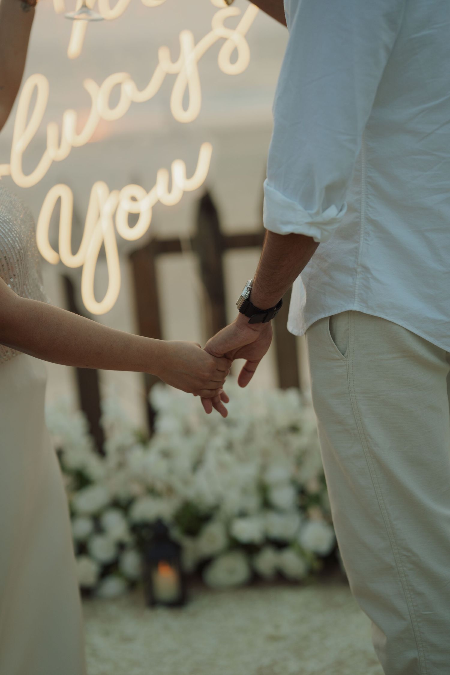 Couple holding hands at a romantic wedding ceremony with a glowing neon sign reading always you in the background.