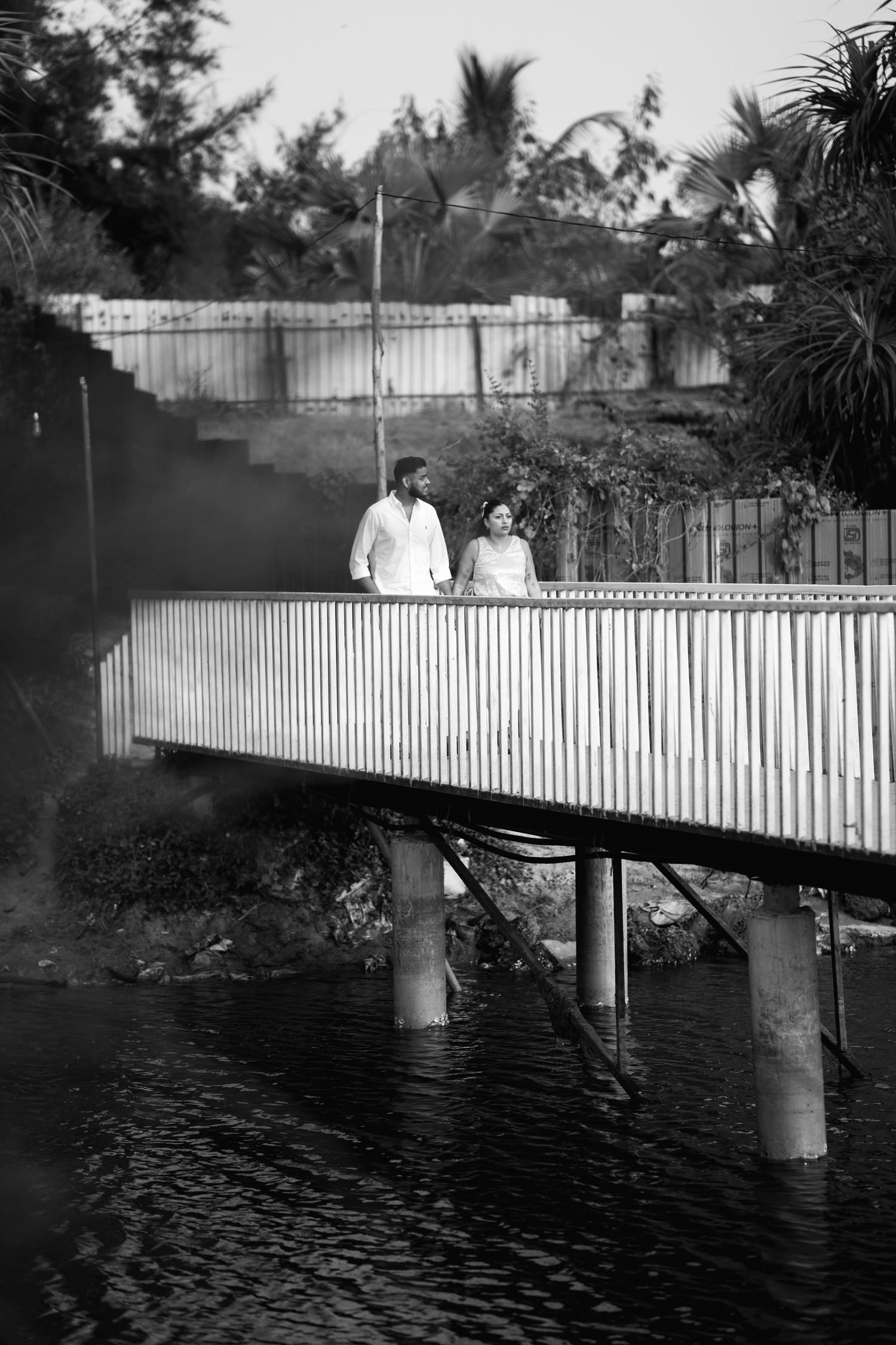 Black and white photo of a couple standing on a modern bridge over water, surrounded by lush tropical greenery.