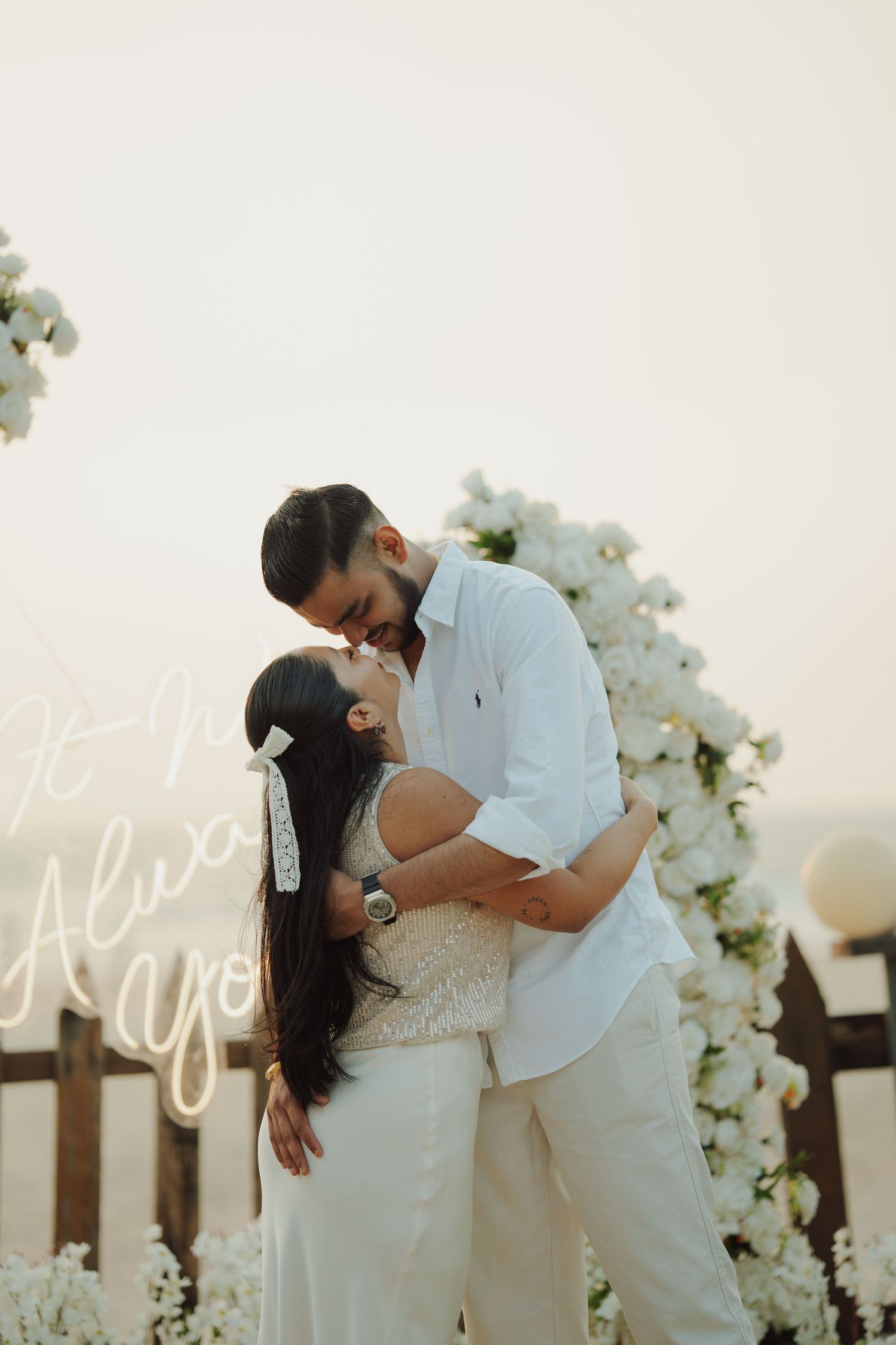 Couple sharing a romantic kiss at an outdoor engagement setup with white floral arch and neon sign at sunset.