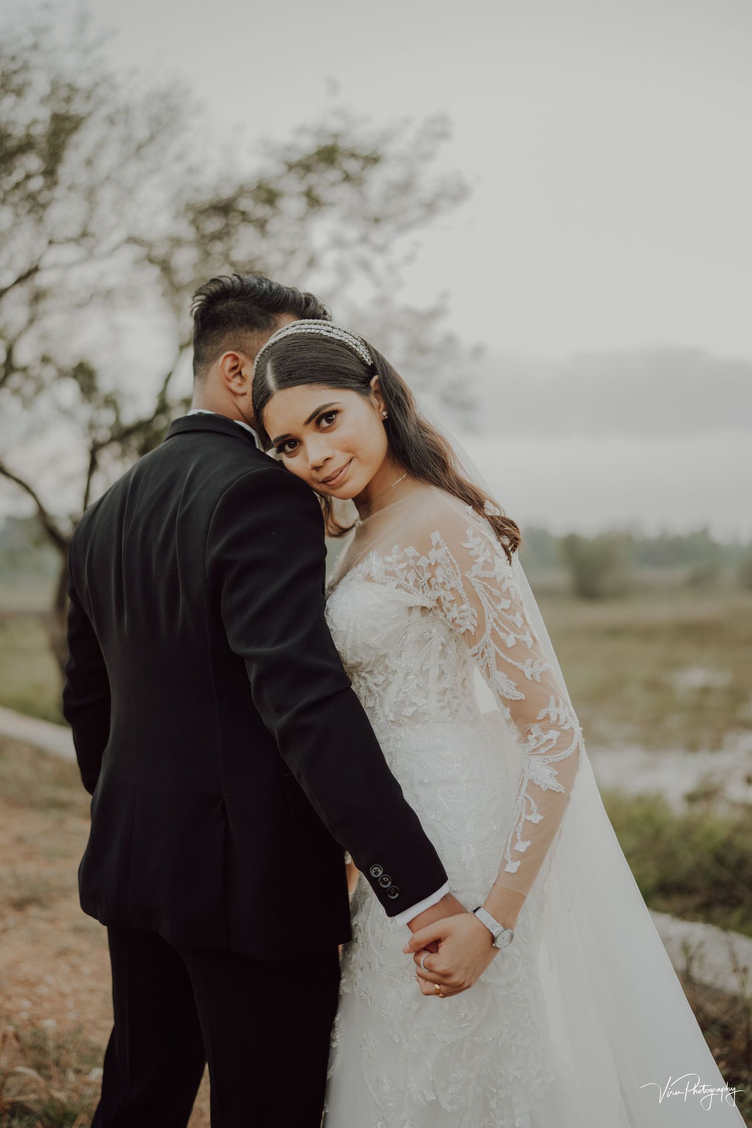 Newlyweds pose for romantic outdoor wedding photo in misty natural setting while bride wears lace sleeve gown.