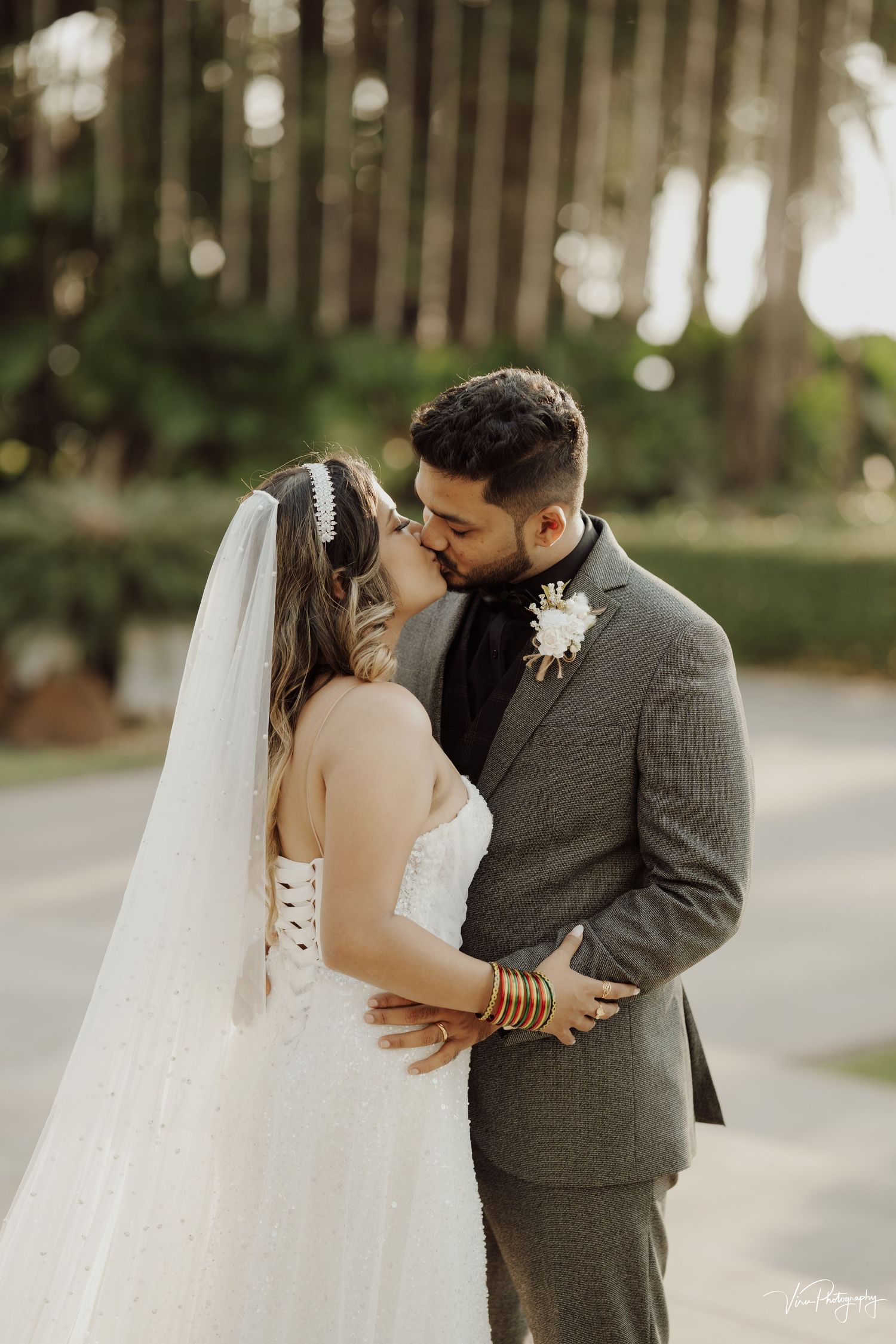 A romantic wedding couple embraces outdoors with the bride in white and groom in gray suit surrounded by trees.