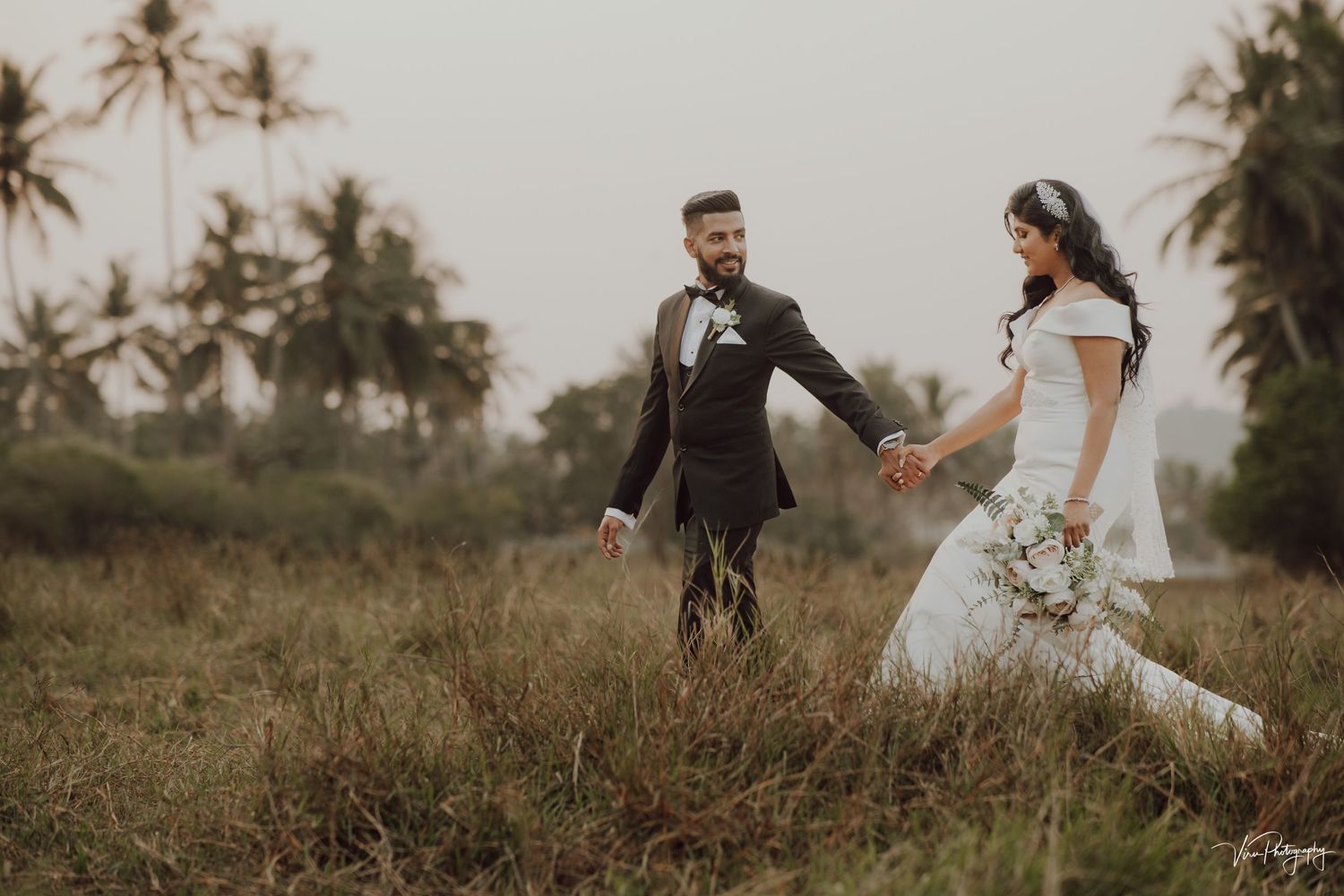 A couple in wedding attire walks hand in hand through a tropical field with palm trees at sunset.