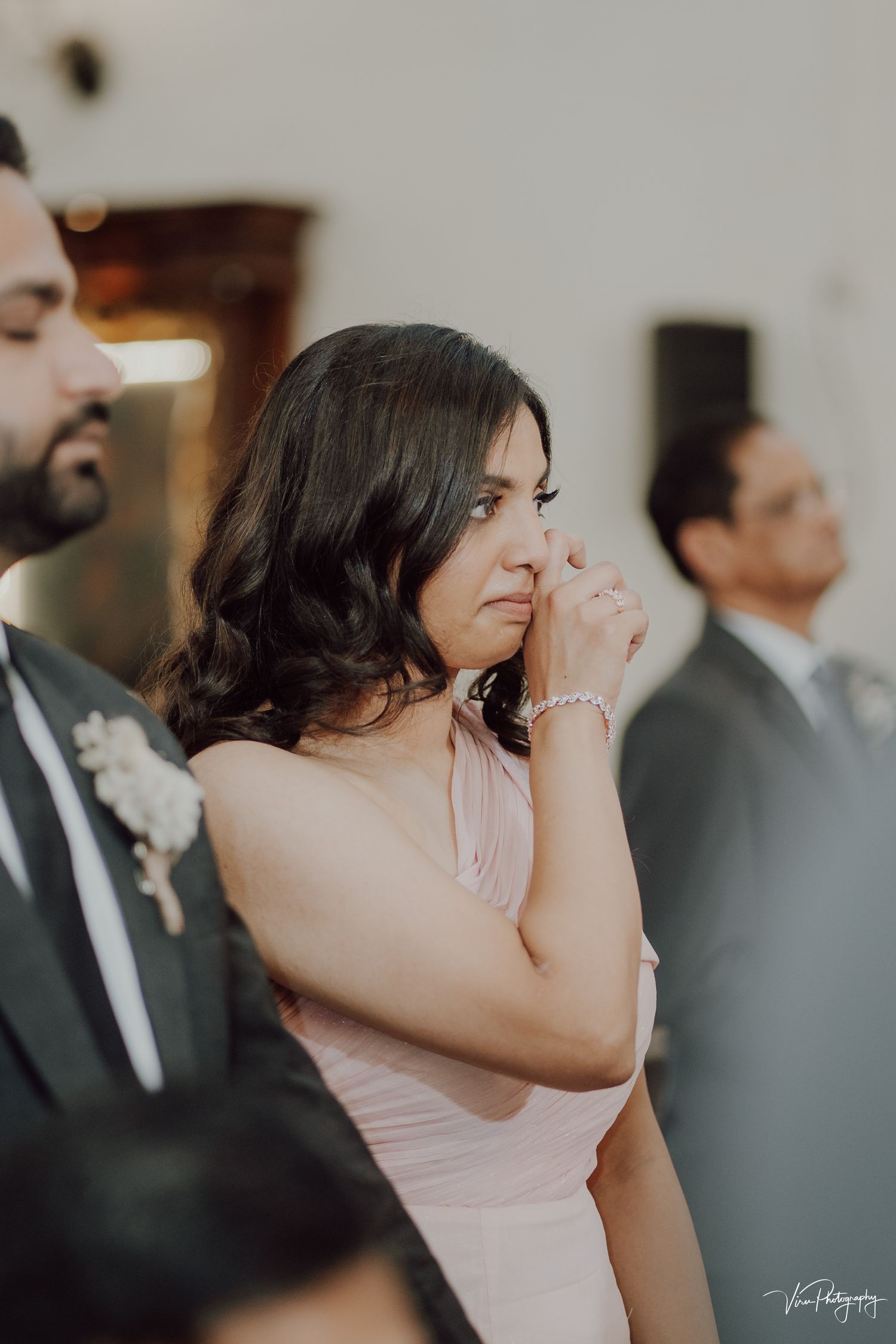 An emotional guest in a pink dress wipes away tears during what appears to be a wedding ceremony.