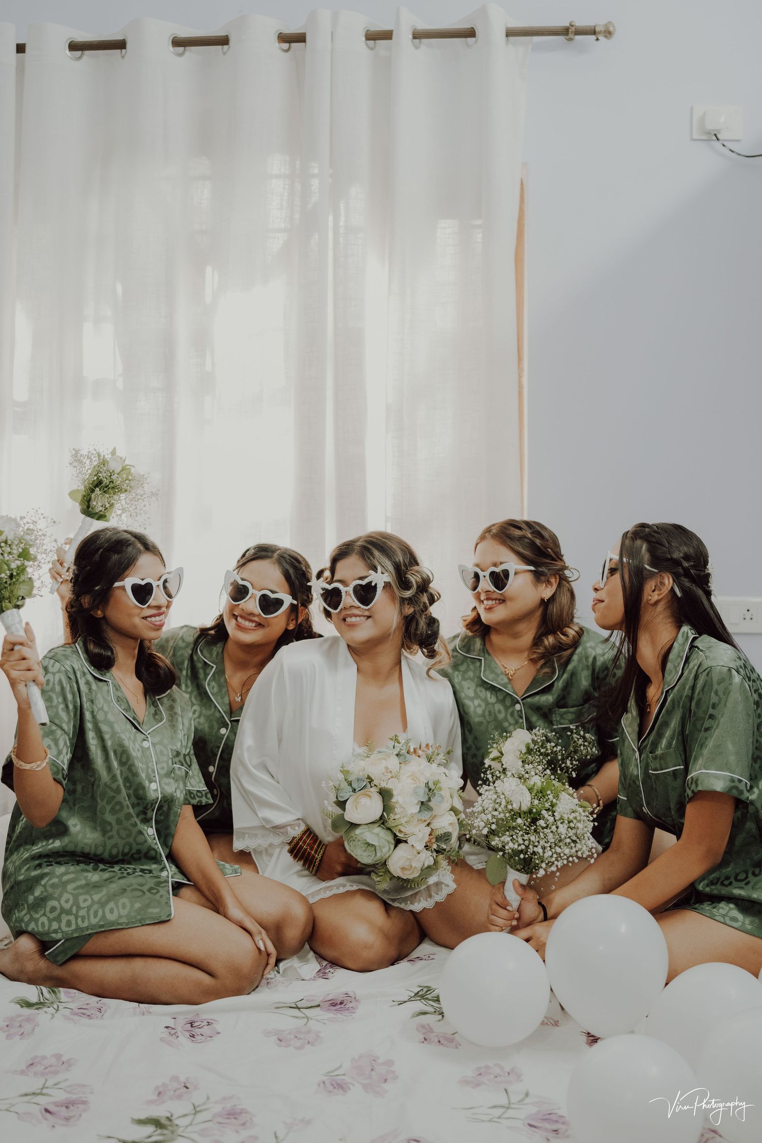 A group in green dresses and sunglasses poses together with white balloons and flowers during a pre-wedding celebration.