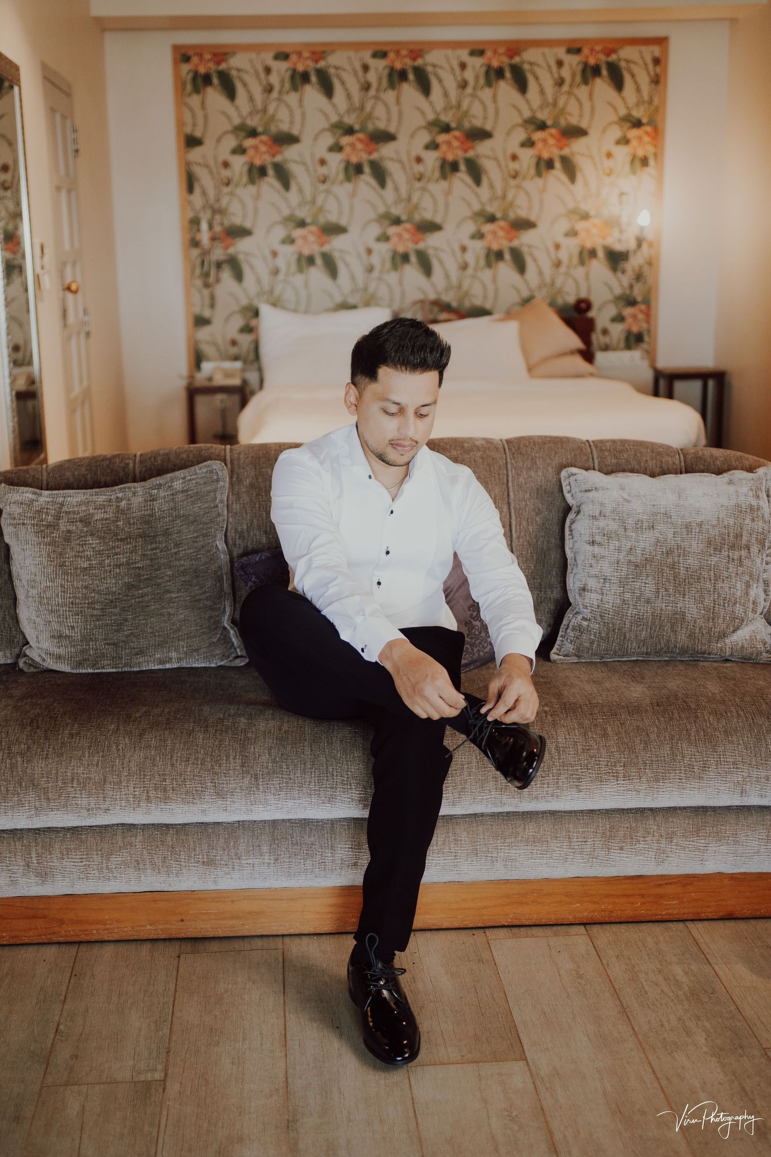 Person in white button-down shirt and dark pants sits on gray couch putting on formal black shoes in bedroom setting.