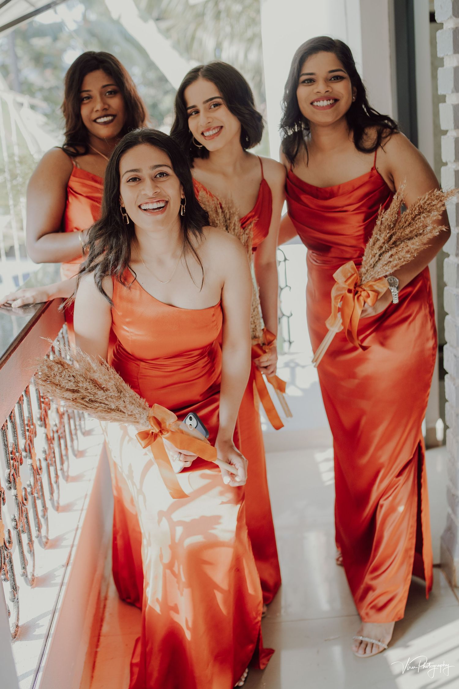 Four bridesmaids in coral silk dresses pose together on a white porch while holding dried pampas grass bouquets.