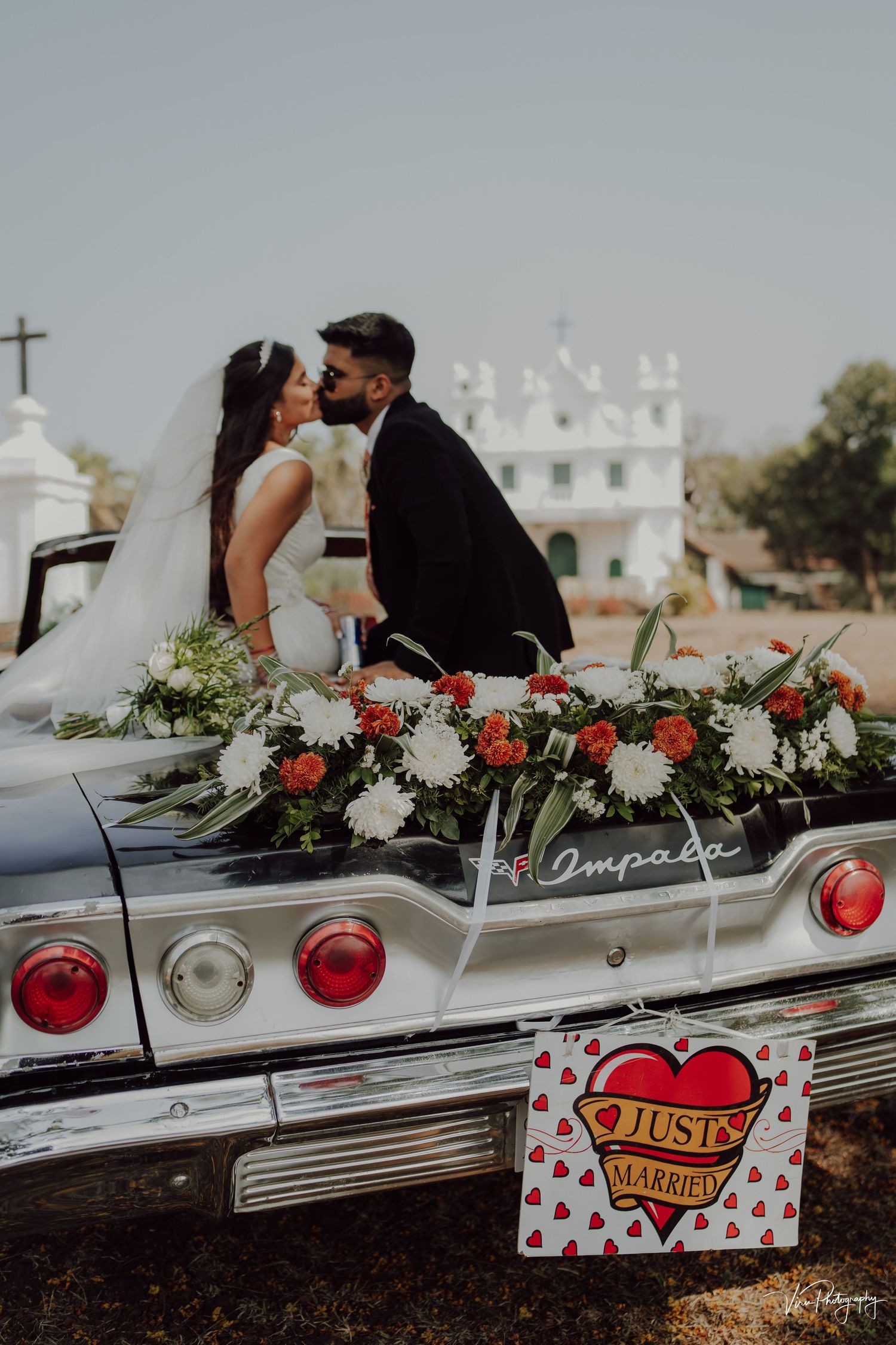 A classic white Impala decorated with red and white flowers serves as backdrop for a wedding photoshoot next to a church.