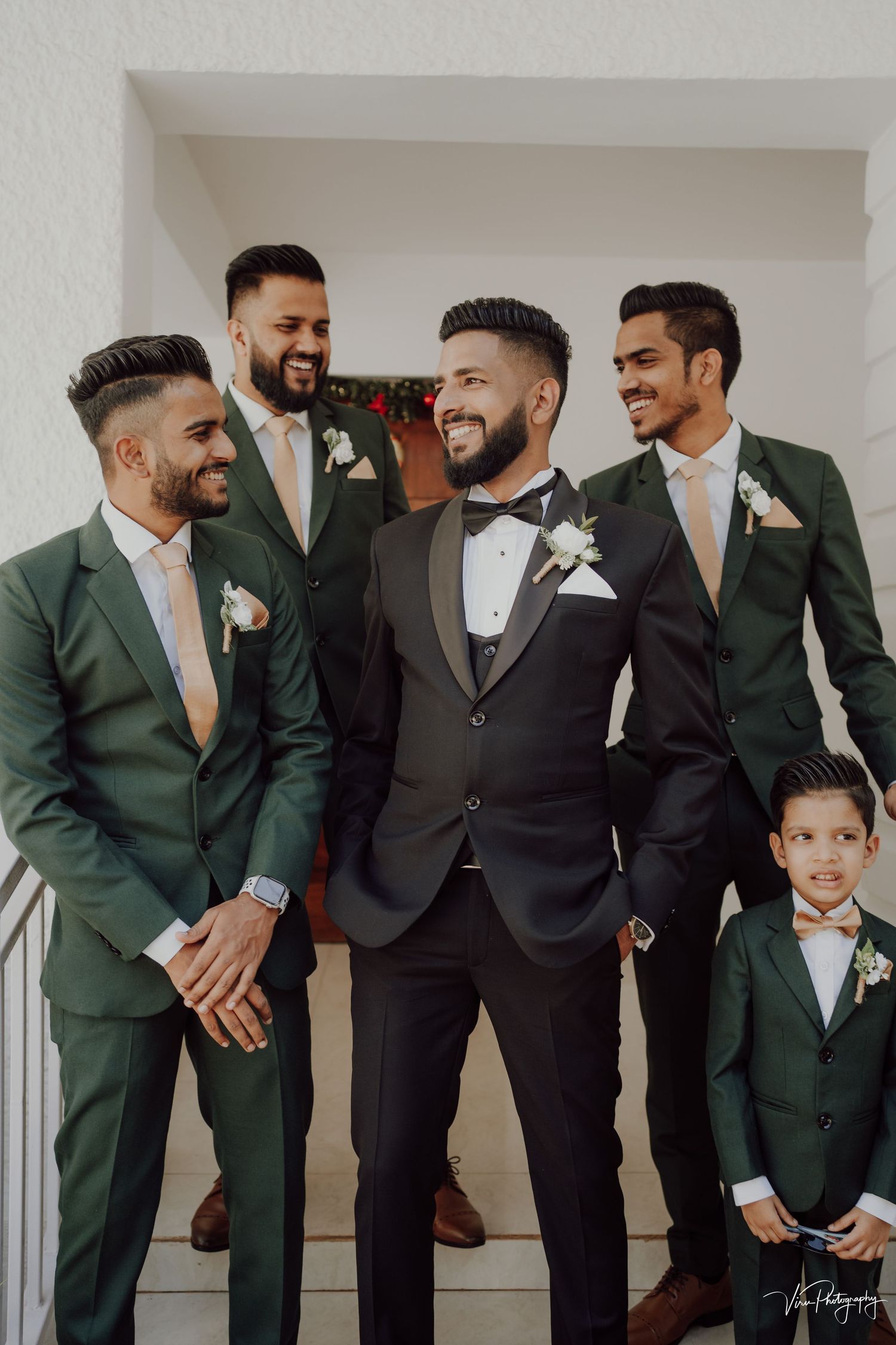 Groomsmen share a laugh while wearing emerald green and charcoal suits with matching boutonnieres at a wedding.