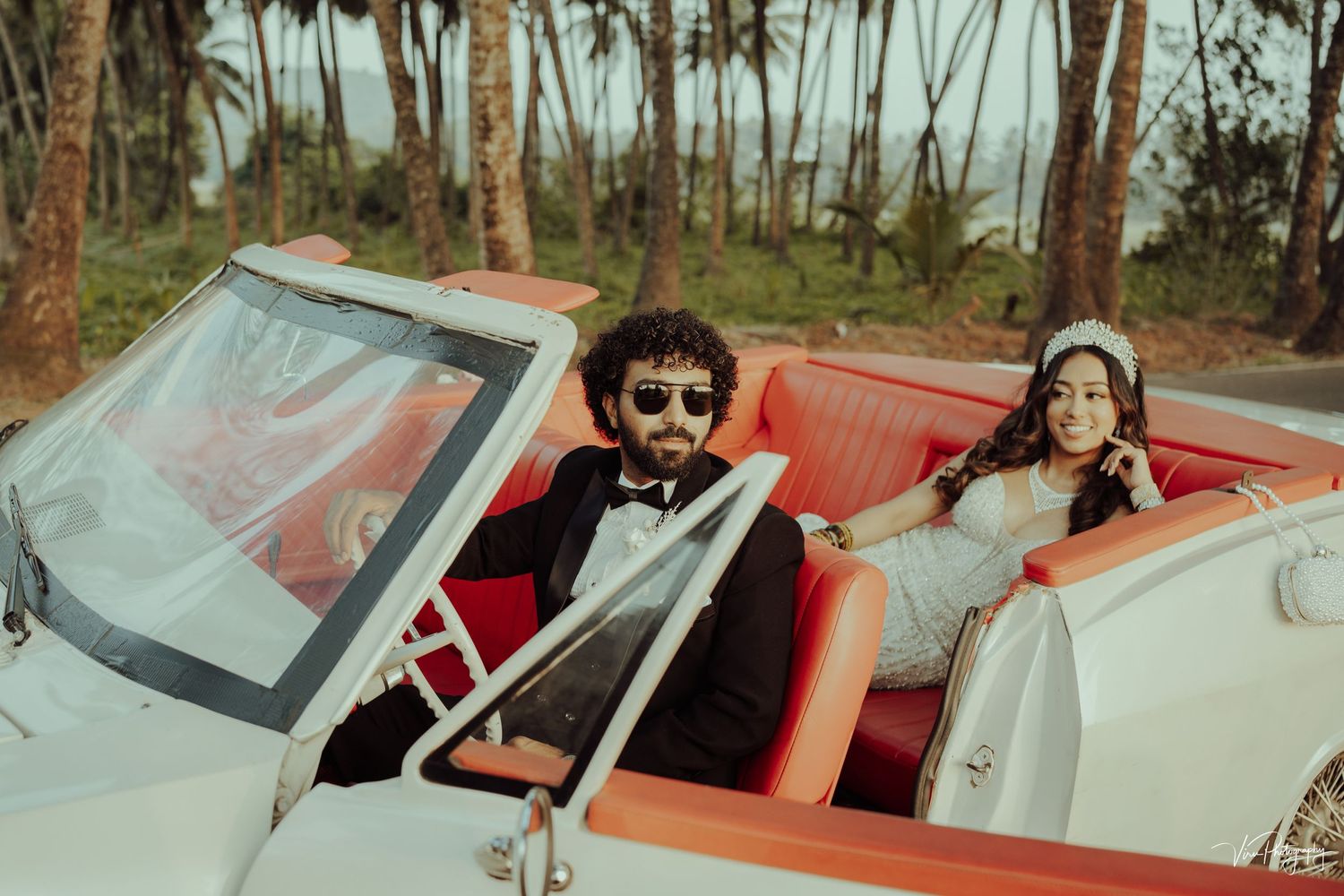 Two people cruising in a vintage white convertible car with red interior seats on a sunny day with palm trees in background.
