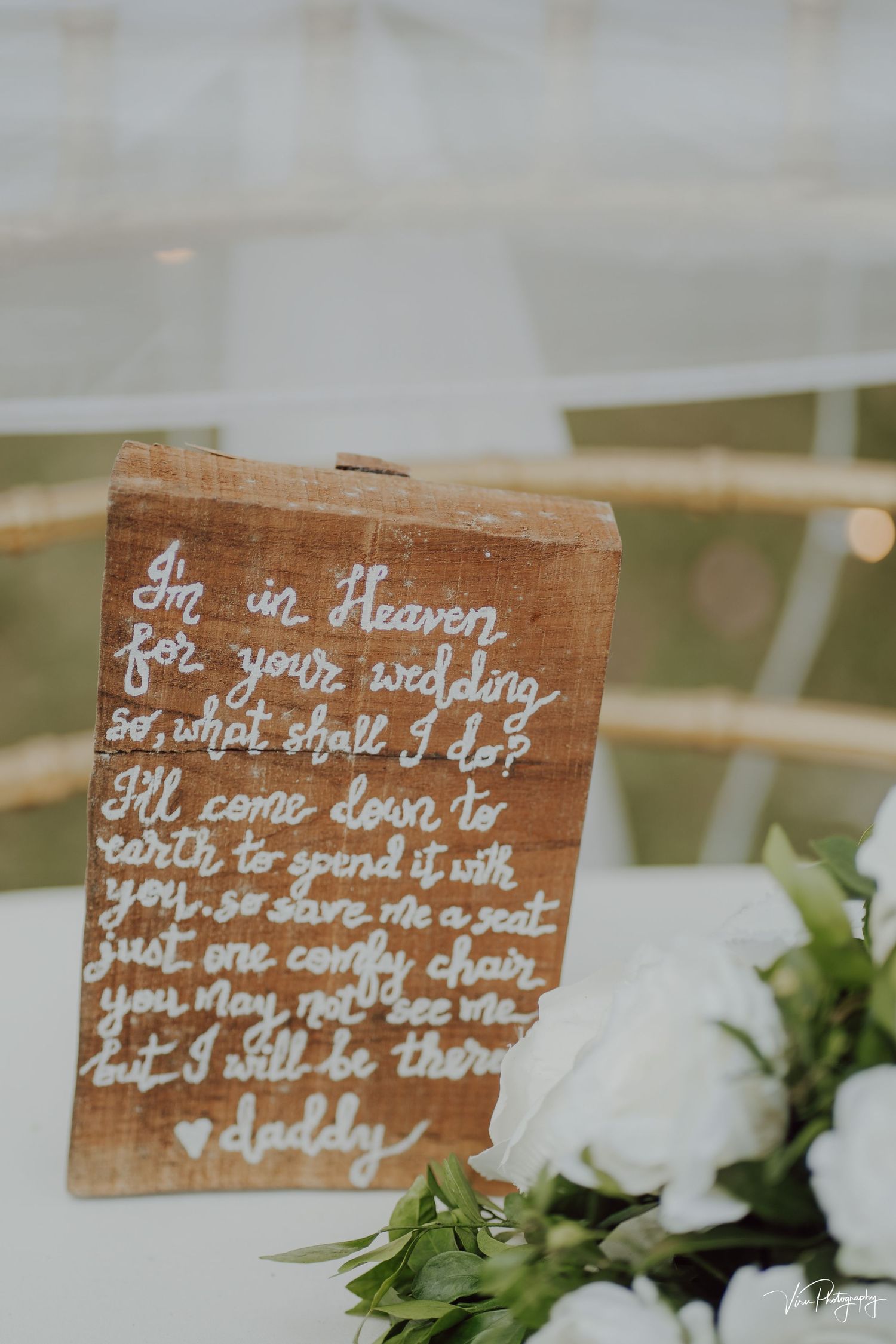 A handwritten message on rustic wood about heaven and weddings sits next to white floral arrangements.
