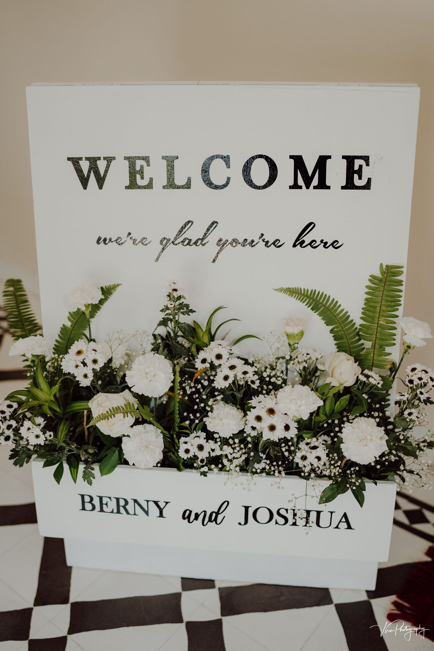 A white welcome sign with black lettering and delicate white floral arrangements on a checkerboard floor.