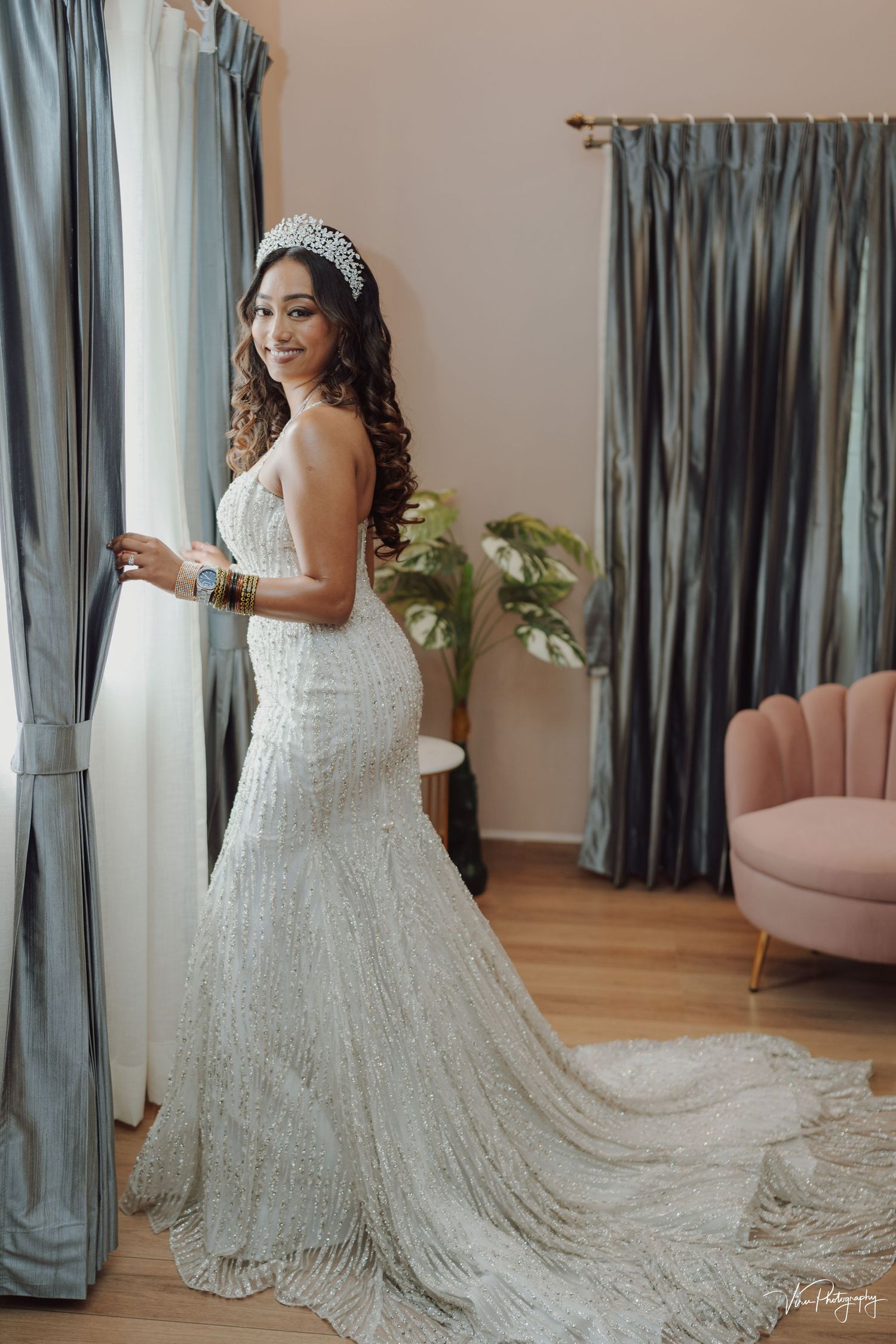 A stunning lace wedding gown with a long train is showcased in an elegant room with blue curtains and pink accent chair.