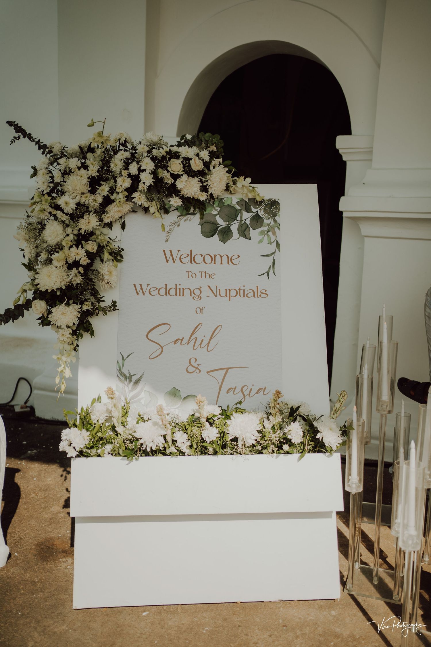 Elegant white wedding welcome sign with floral arrangements and greenery decorating a curved doorway entrance.