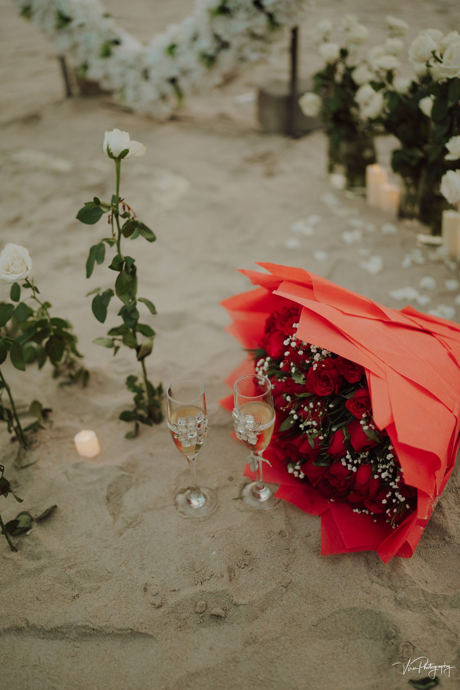 A romantic arrangement with red roses wrapped in coral paper sits on sand next to wine glasses and white floral decor.
