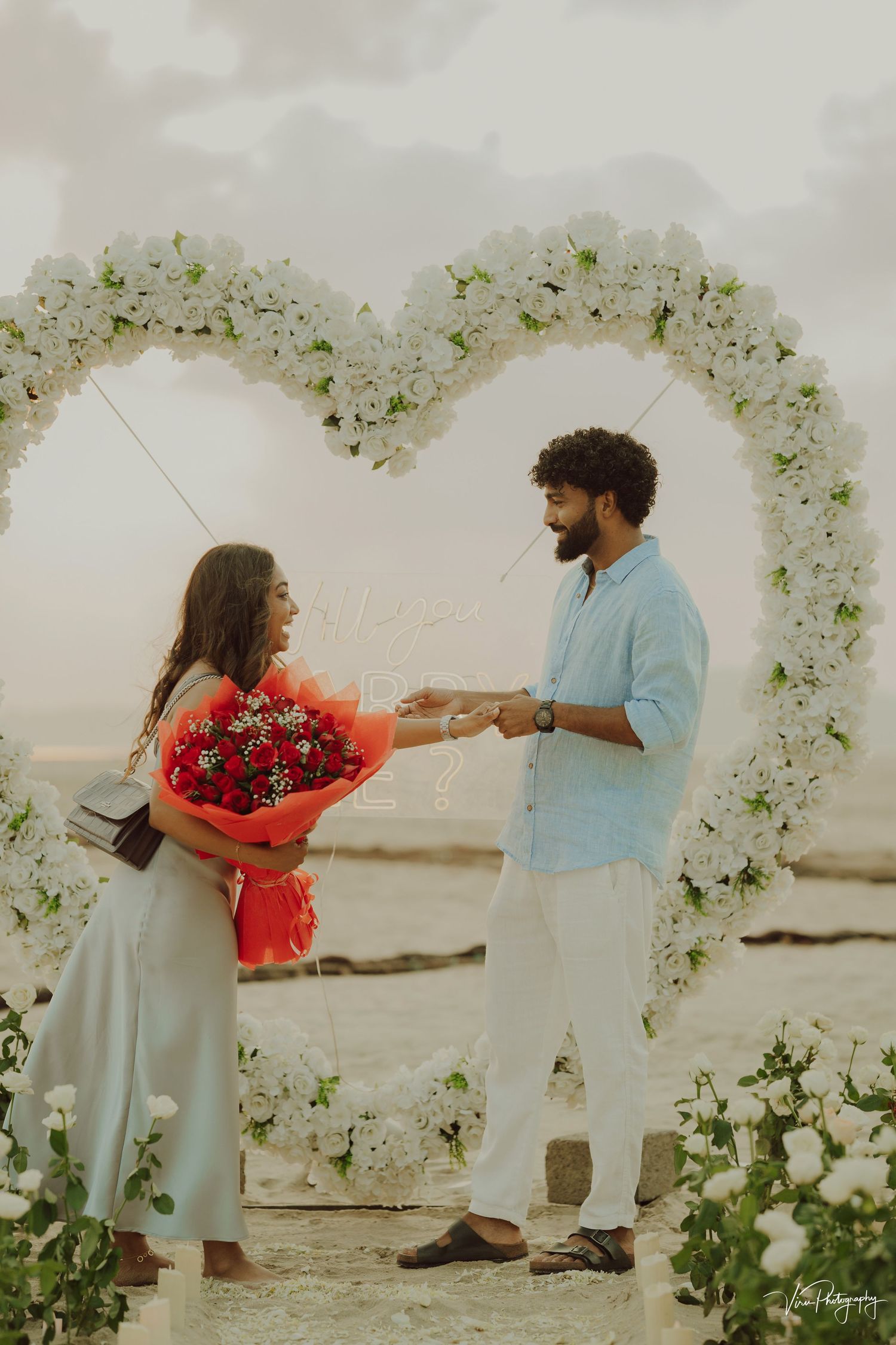 A romantic beach proposal scene with white floral arch decor as a couple shares a moment holding red roses by the ocean.