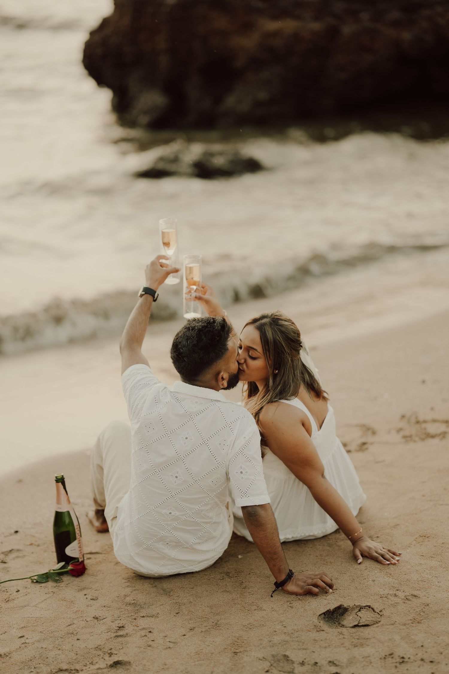 A romantic couple in white clothing laying on the beach sand while sharing a champagne toast at sunset.