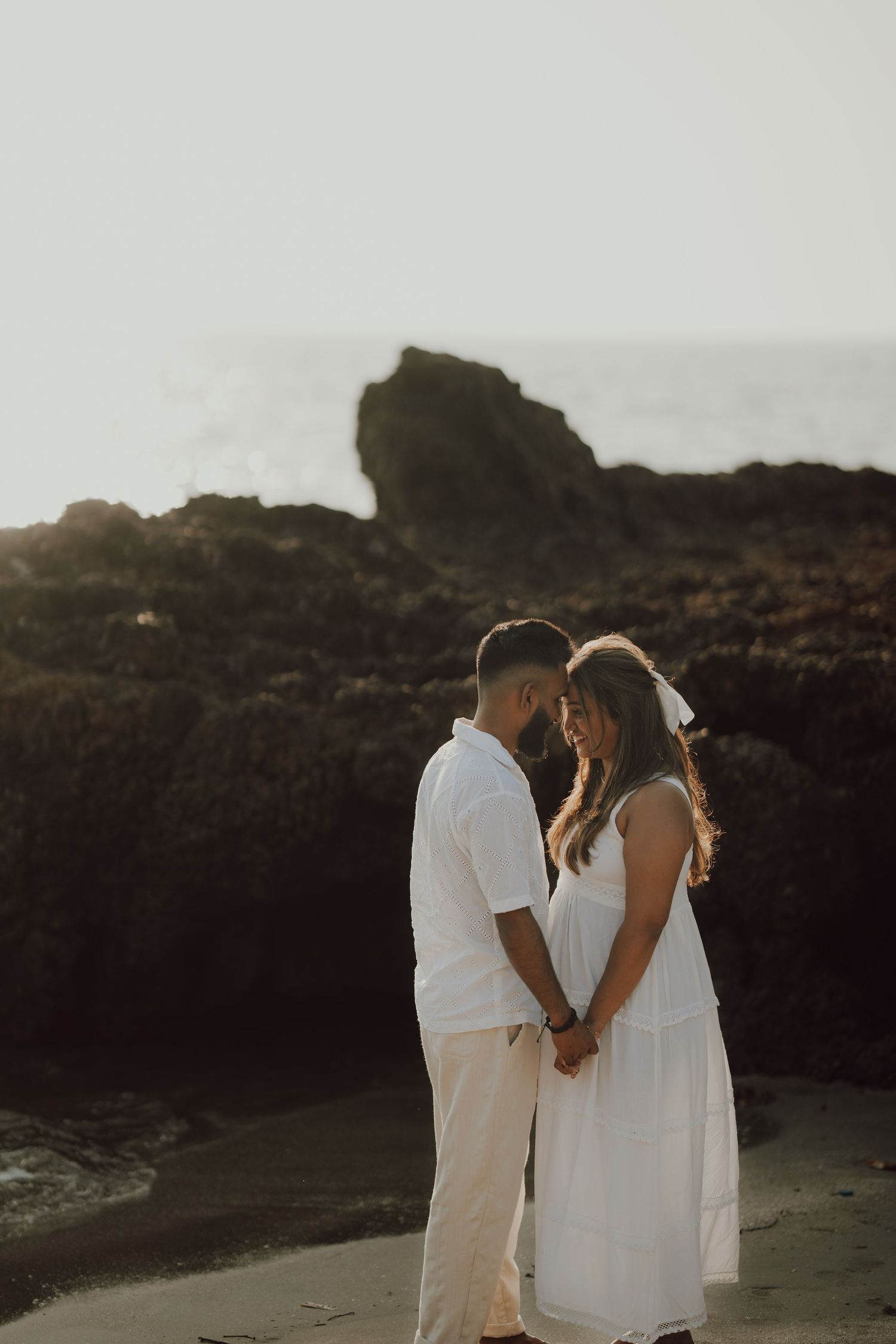 A romantic couple in white outfits embraces on a rocky beach at sunset, creating a dreamy silhouette against the cliffs.