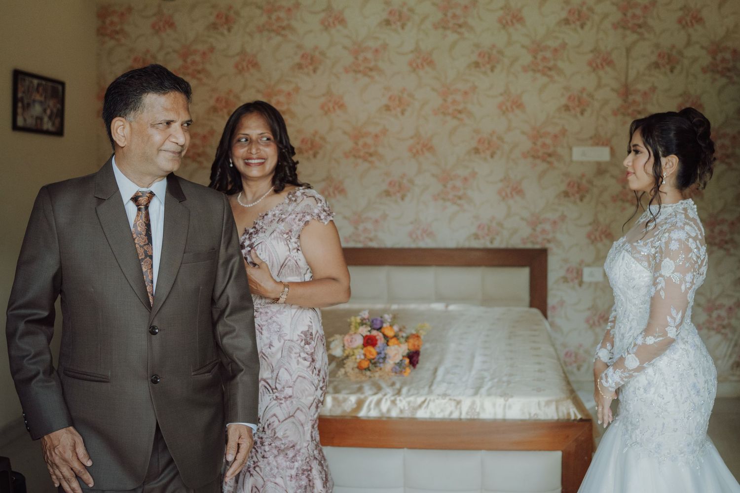 Bride in a white lace gown stands facing her parents, a man in a brown suit and a woman in a floral dress, in a warmly lit bedroom.