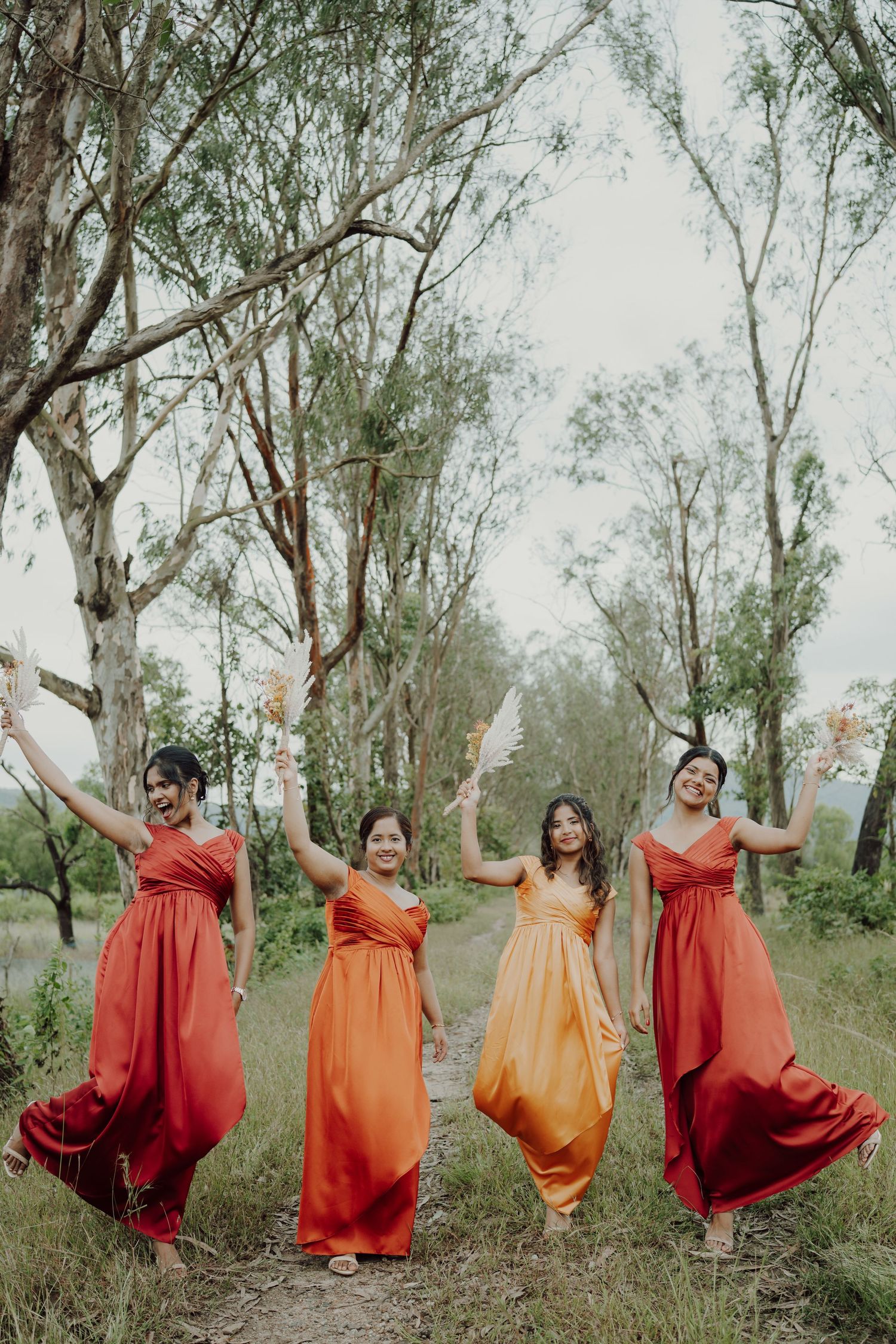 Four bridesmaids in orange and red dresses dance joyfully on a grassy path among tall eucalyptus trees.
