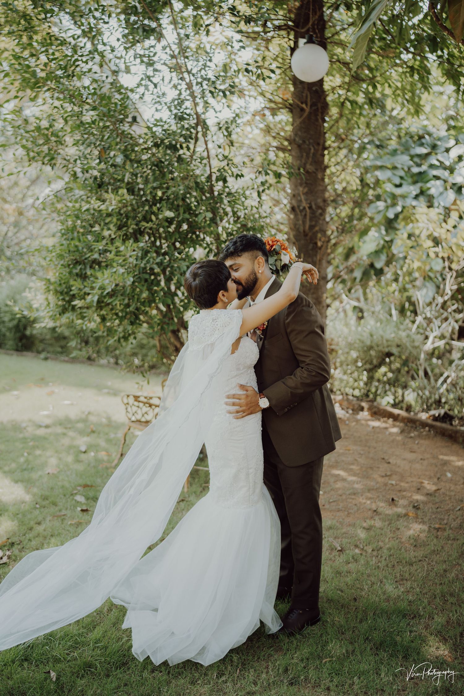 A couple shares an intimate moment in a garden setting with flowing white wedding dress and brown suit.