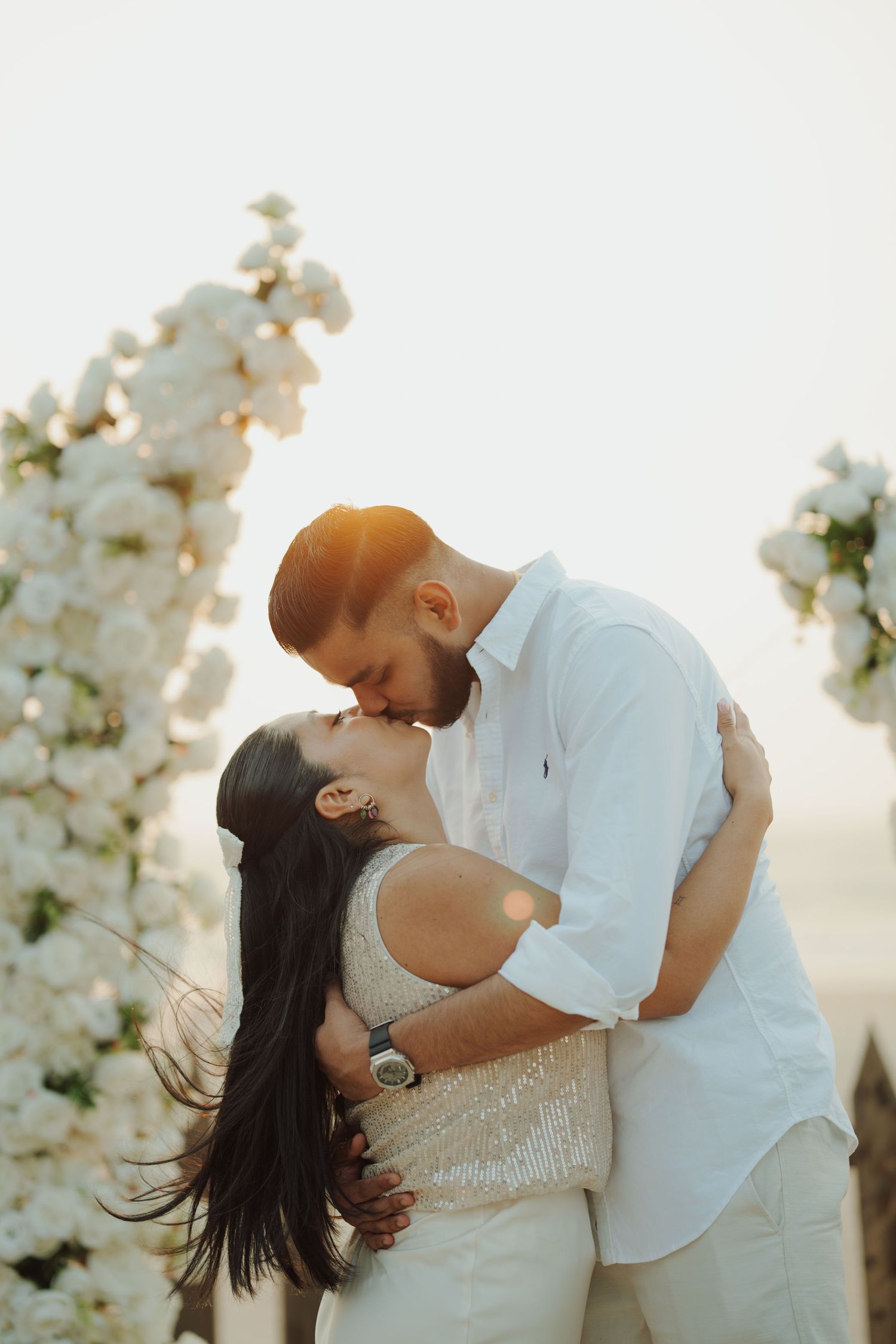 Couple sharing a romantic kiss at golden hour, framed by white floral arrangements at an outdoor wedding ceremony.