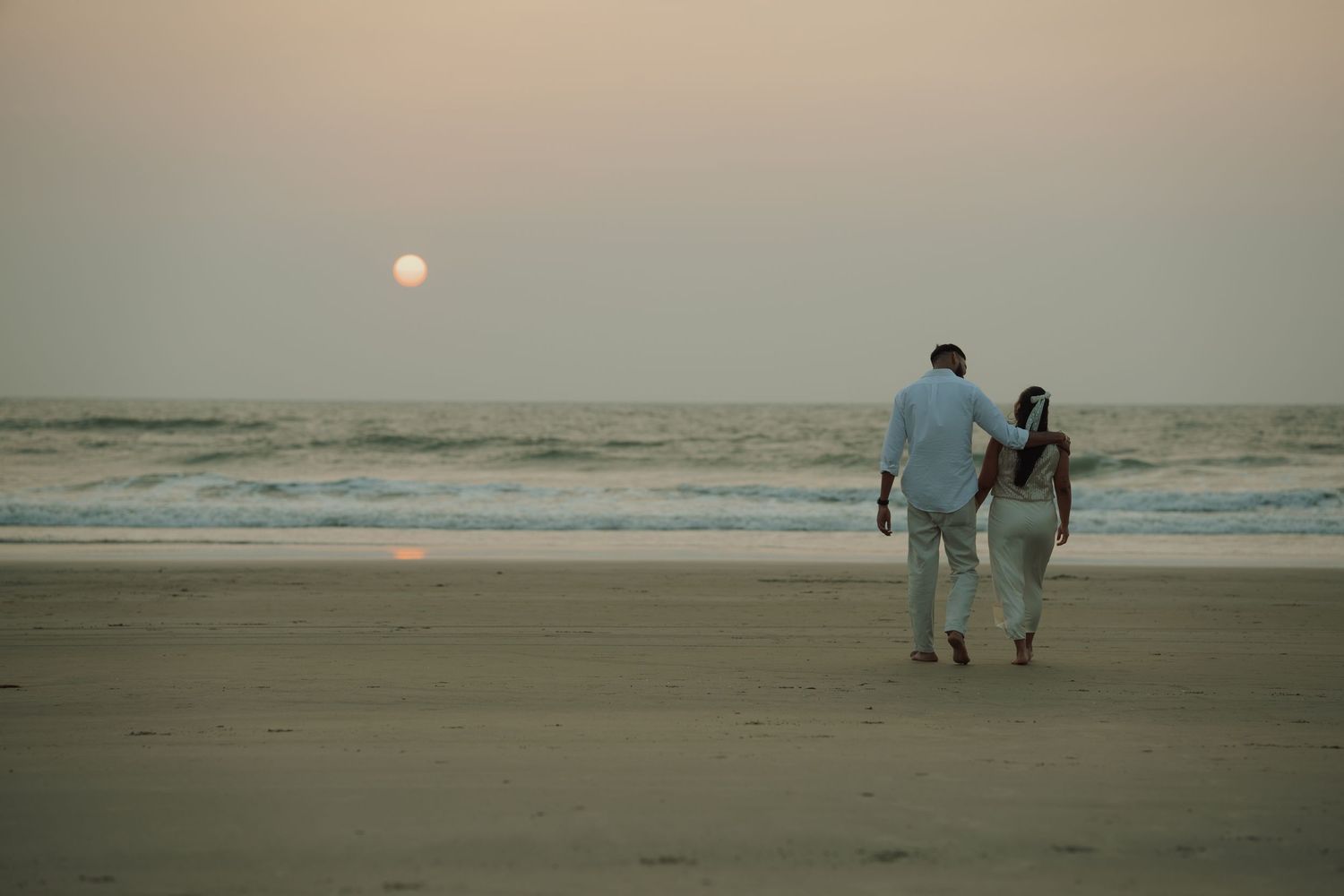 Couple walking hand in hand on a serene beach at sunset with waves and glowing sun on the horizon.
