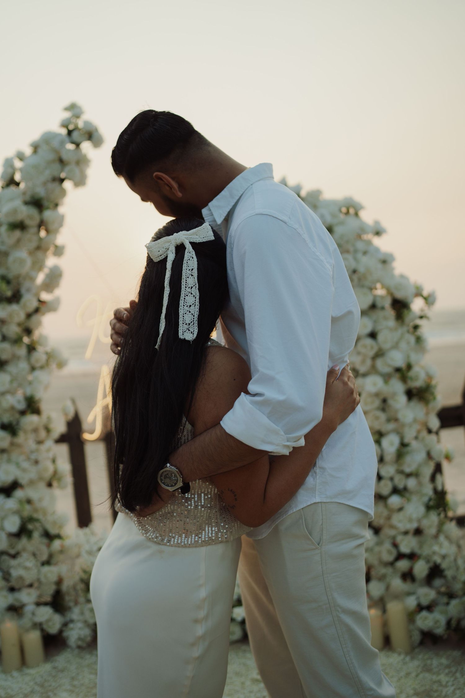 Bride and groom embracing at golden hour before white floral arch at romantic beach wedding ceremony.