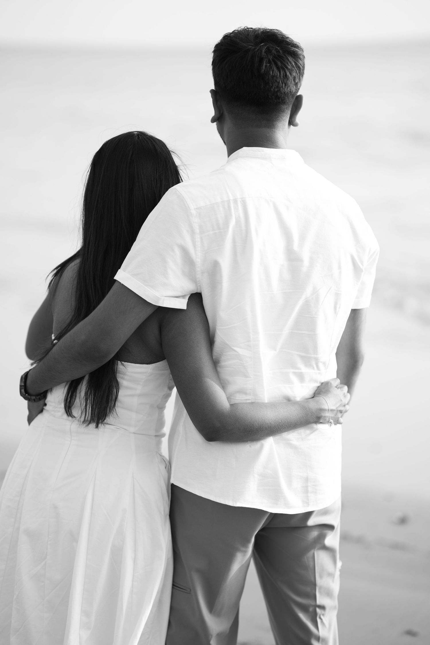 A couple in white clothing embraces while standing on a beach, viewed from behind in black and white photography.