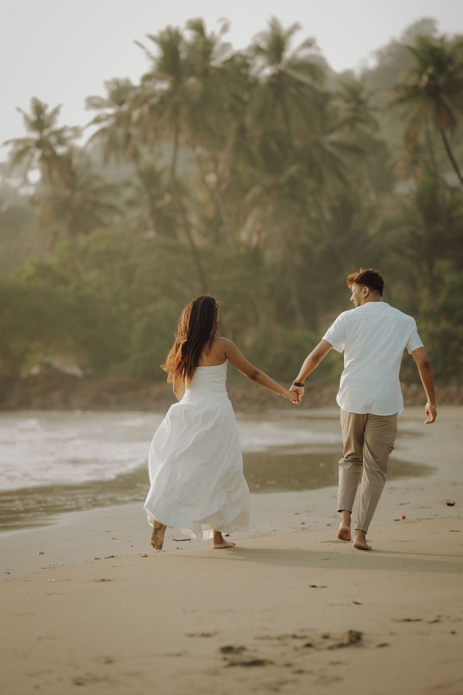 A couple walks playfully along a tropical beach with palm trees at sunset creating a romantic silhouette.