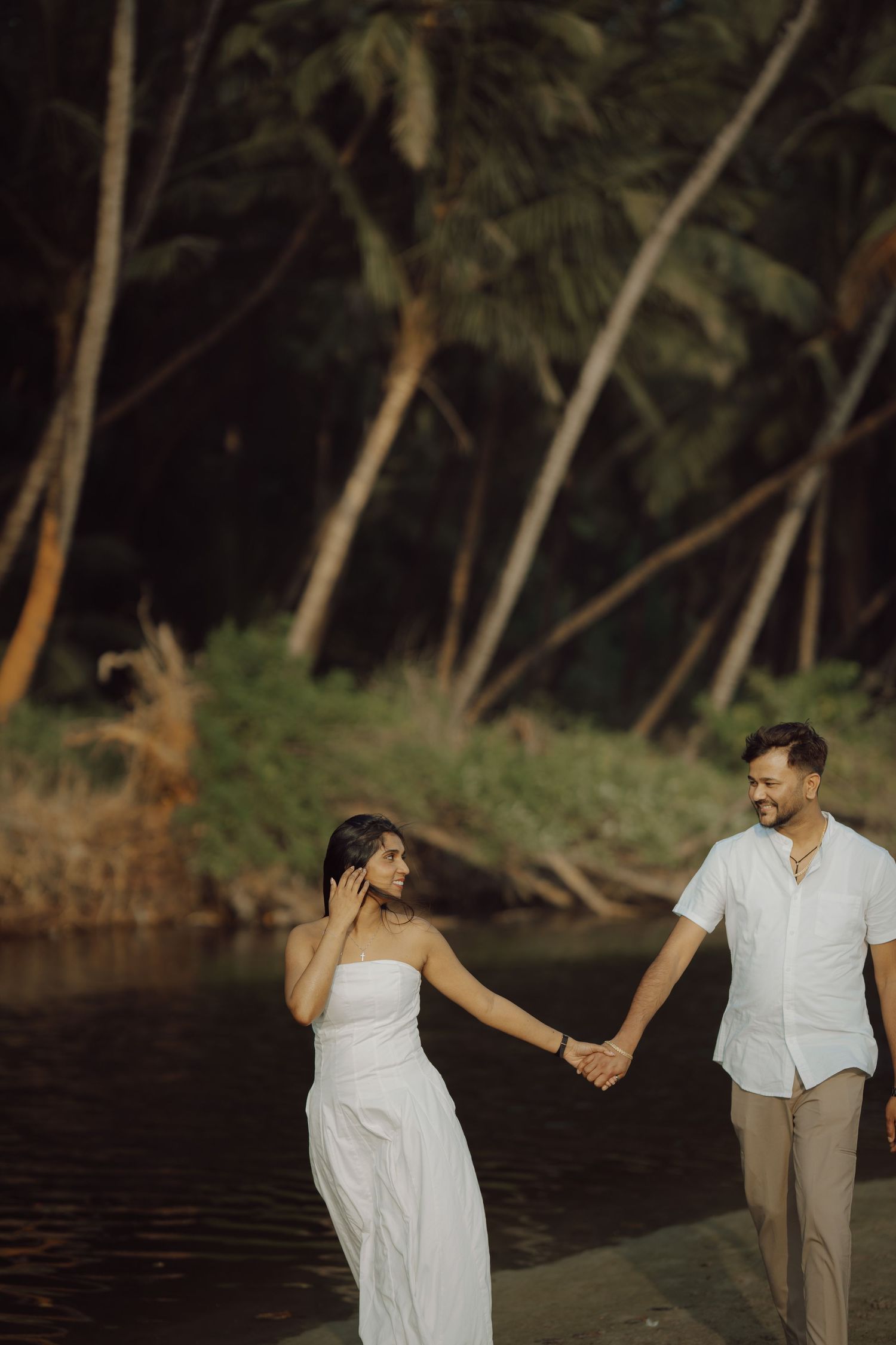 Couple in white attire holding hands while walking along a tropical riverbank with palm trees in the background.