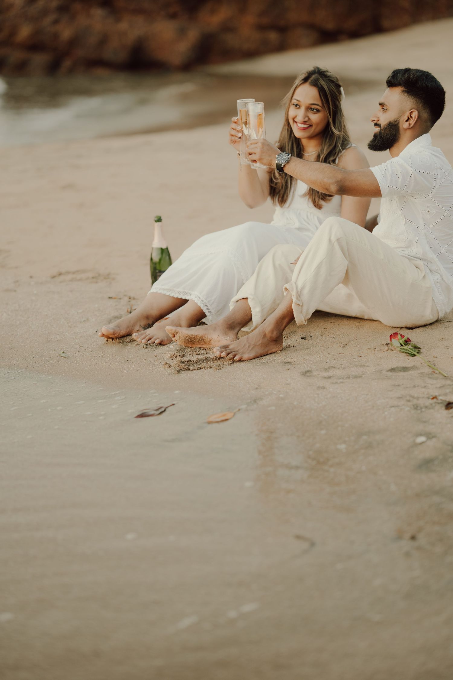 A couple in white attire toasts with champagne glasses while sitting barefoot on a sandy beach at sunset.