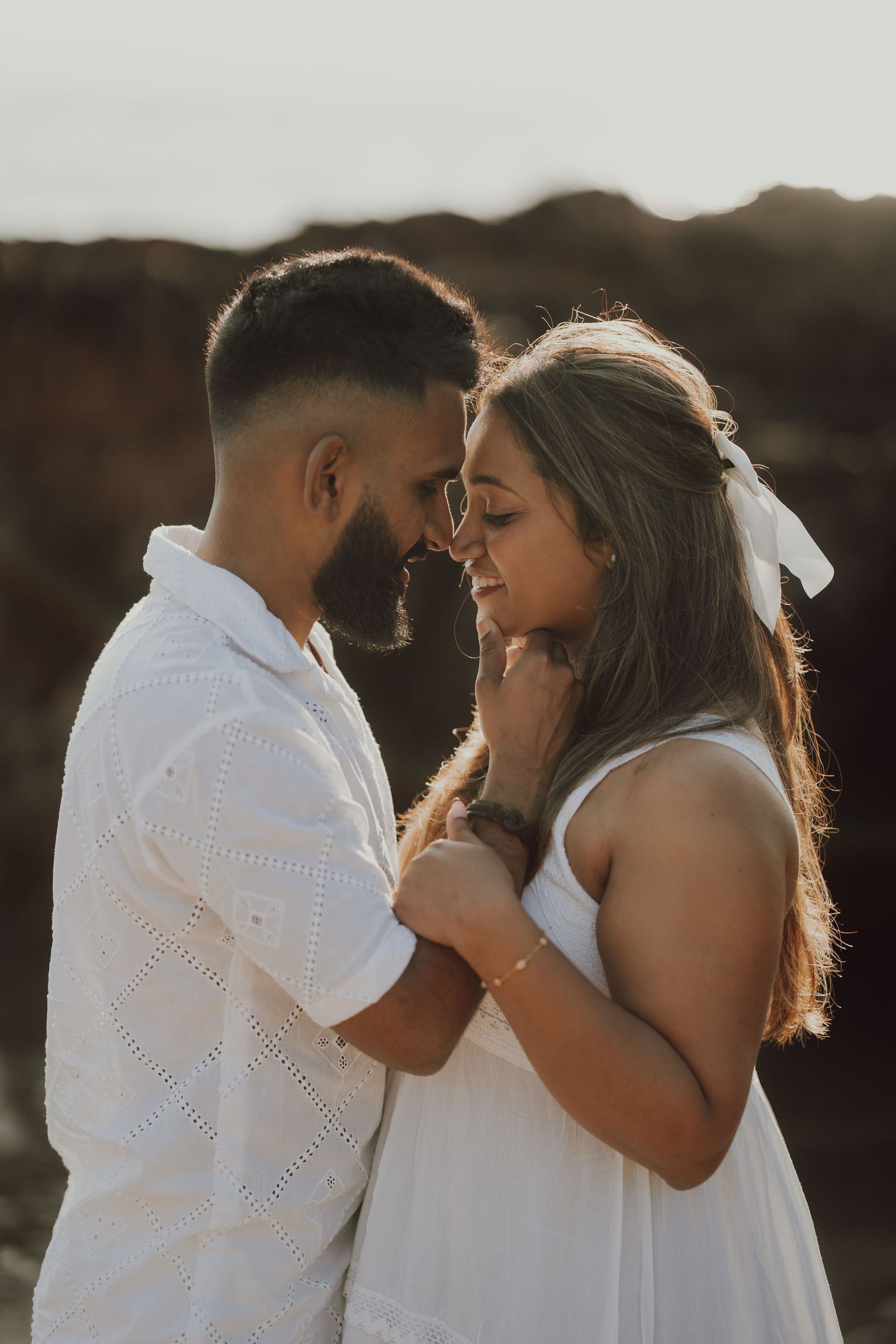 A romantic couple in white outfits share an intimate moment at sunset with golden backlighting.