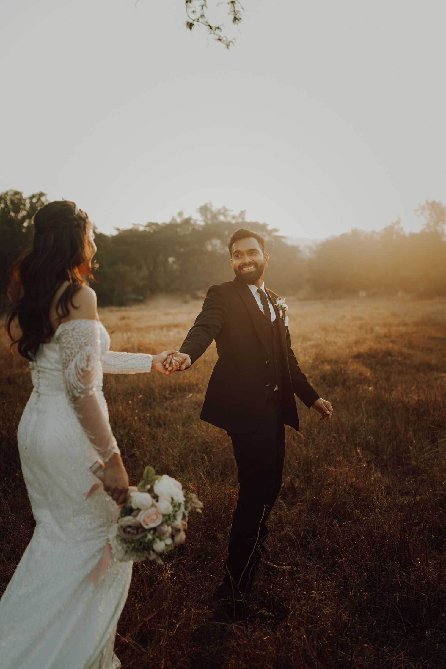 A romantic wedding couple holds hands while walking through a sunset-lit field with golden light.