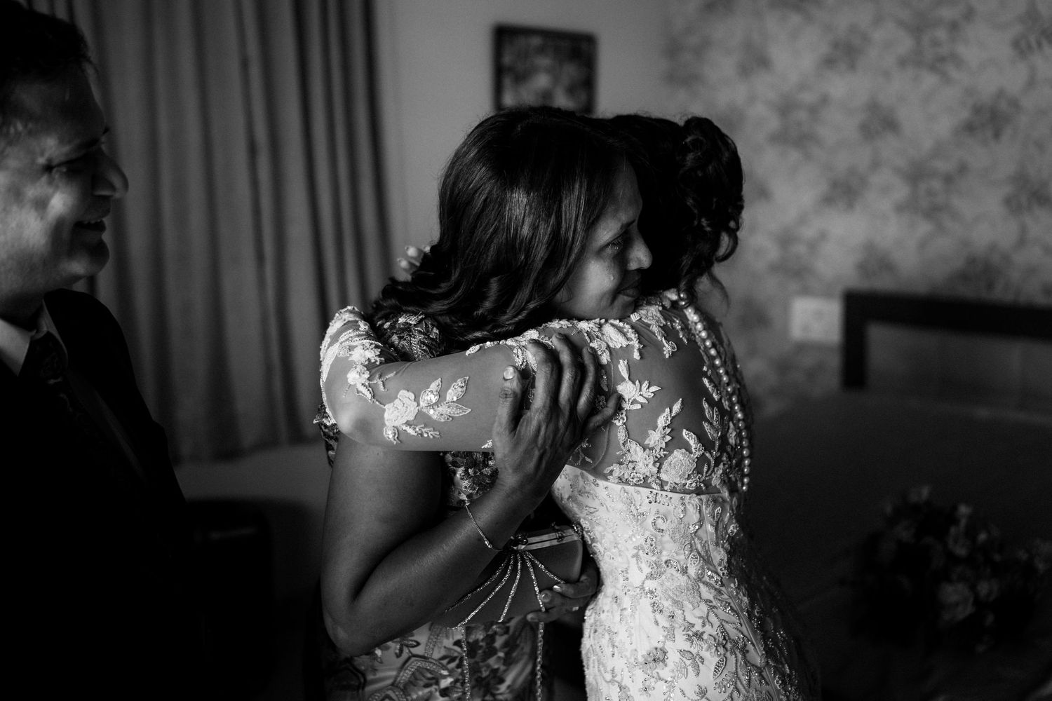 Black and white photo of two women in a tender embrace, one in a lace wedding gown, as a smiling man watches nearby.