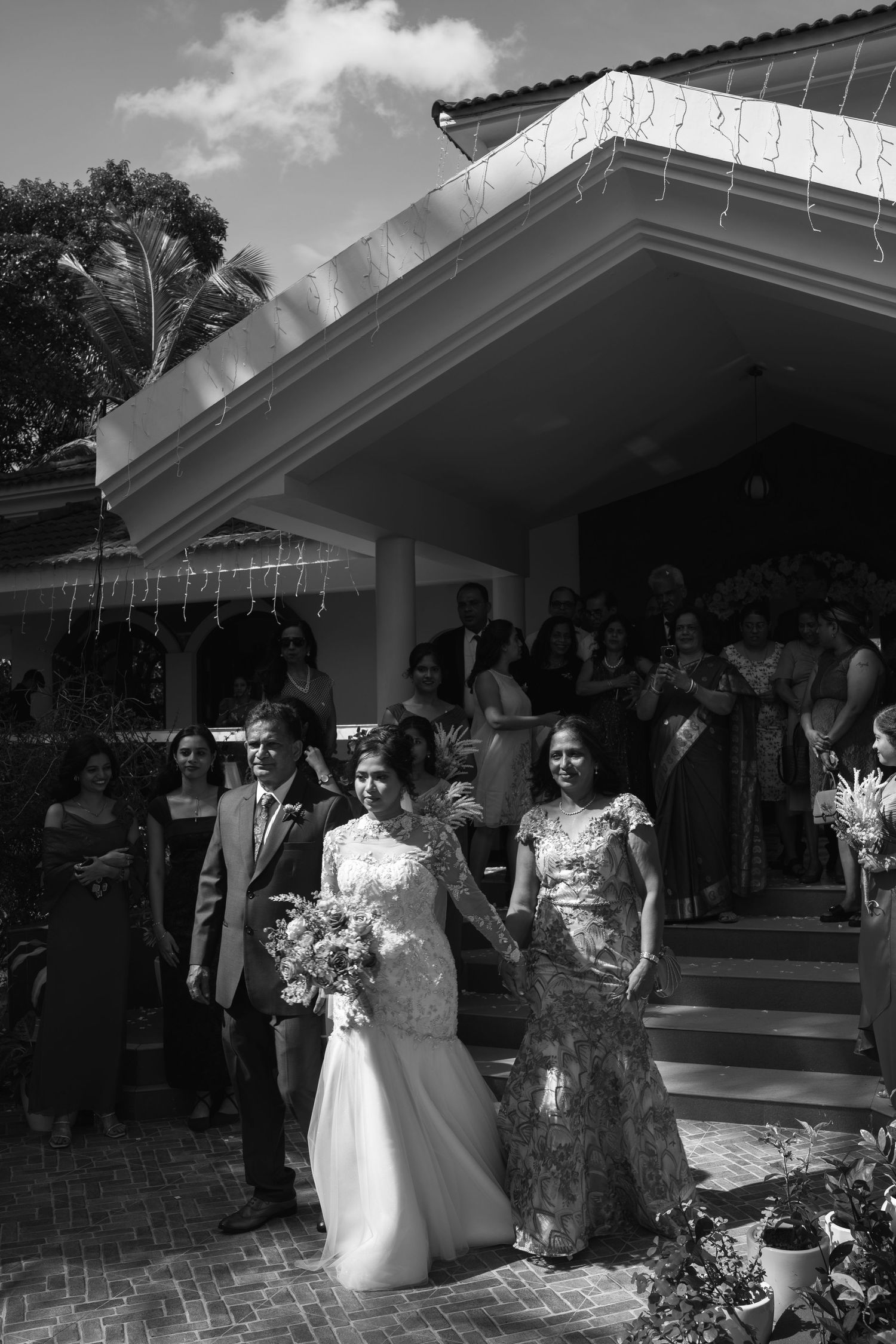 Black and white photo of a bride in a lace gown walking down steps with a woman beside her as wedding guests look on.