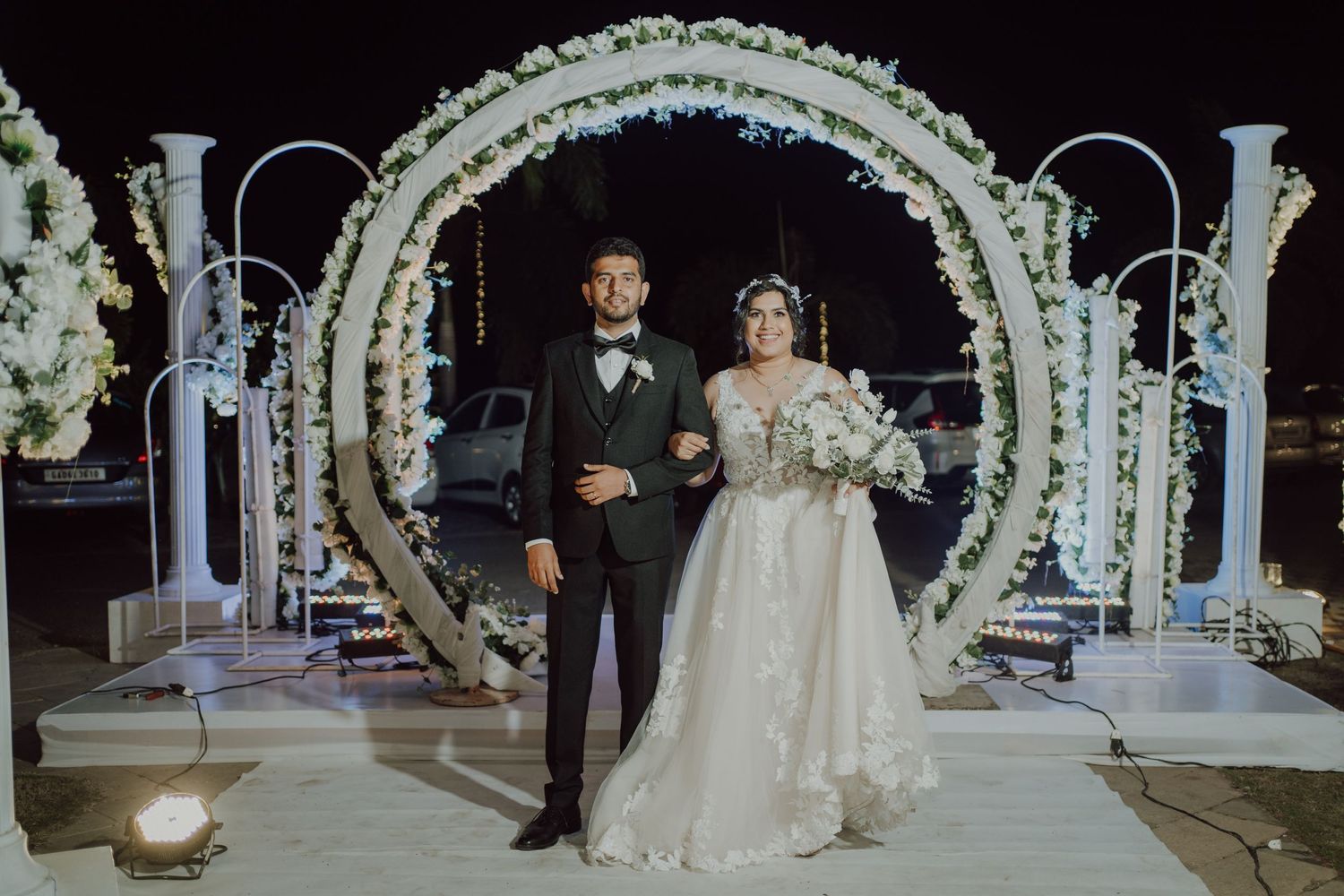 Newlyweds stand under an illuminated floral arch with white flowers and twinkling lights.