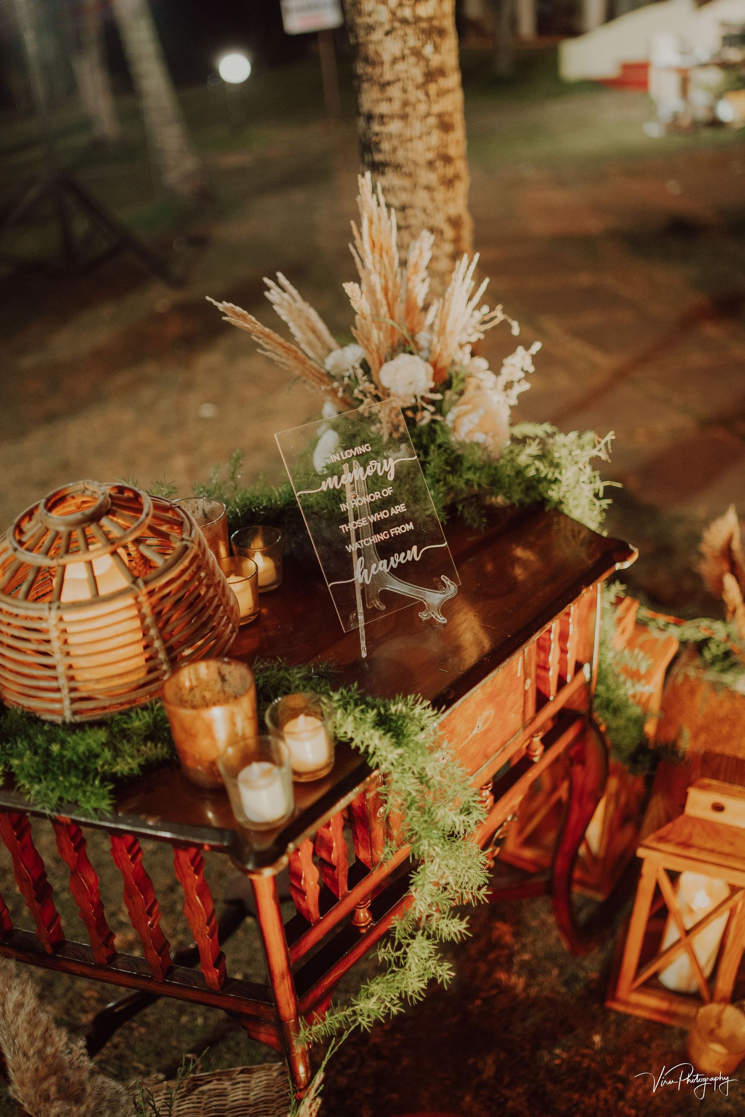 Rustic wedding decor featuring rattan lanterns, wooden crates, dried flowers and glowing candles on an outdoor table at dusk.