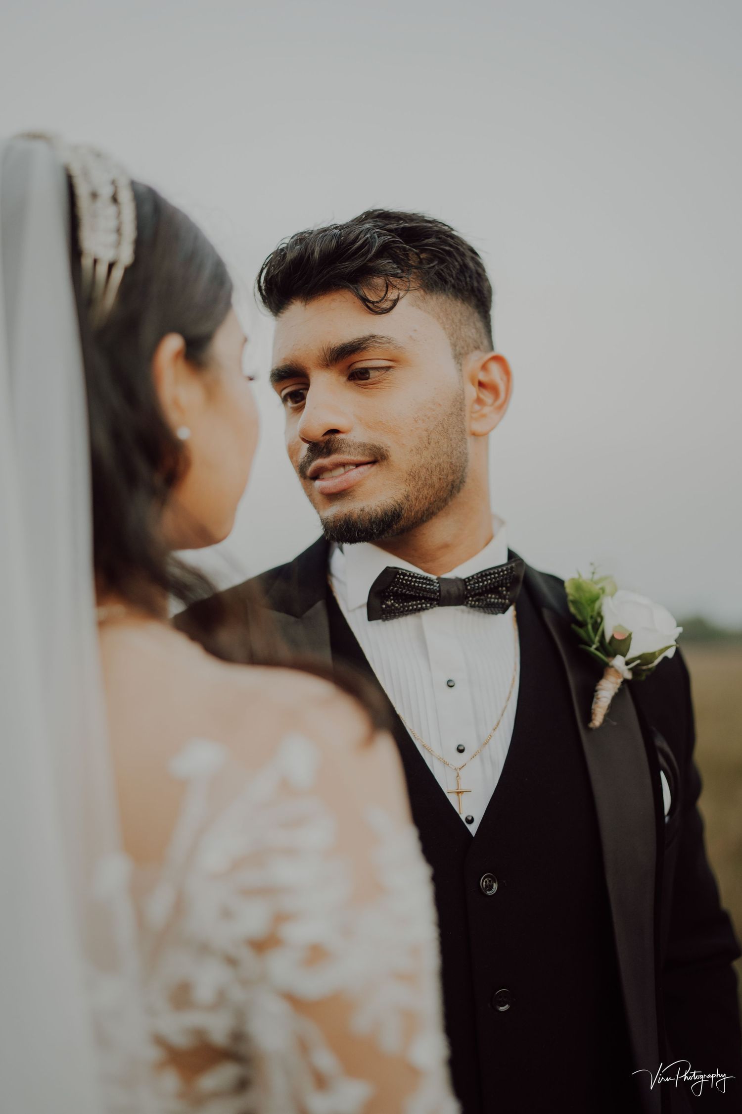 A wedding couple shares an intimate moment as the groom in a black tuxedo gazes lovingly at his veiled bride.