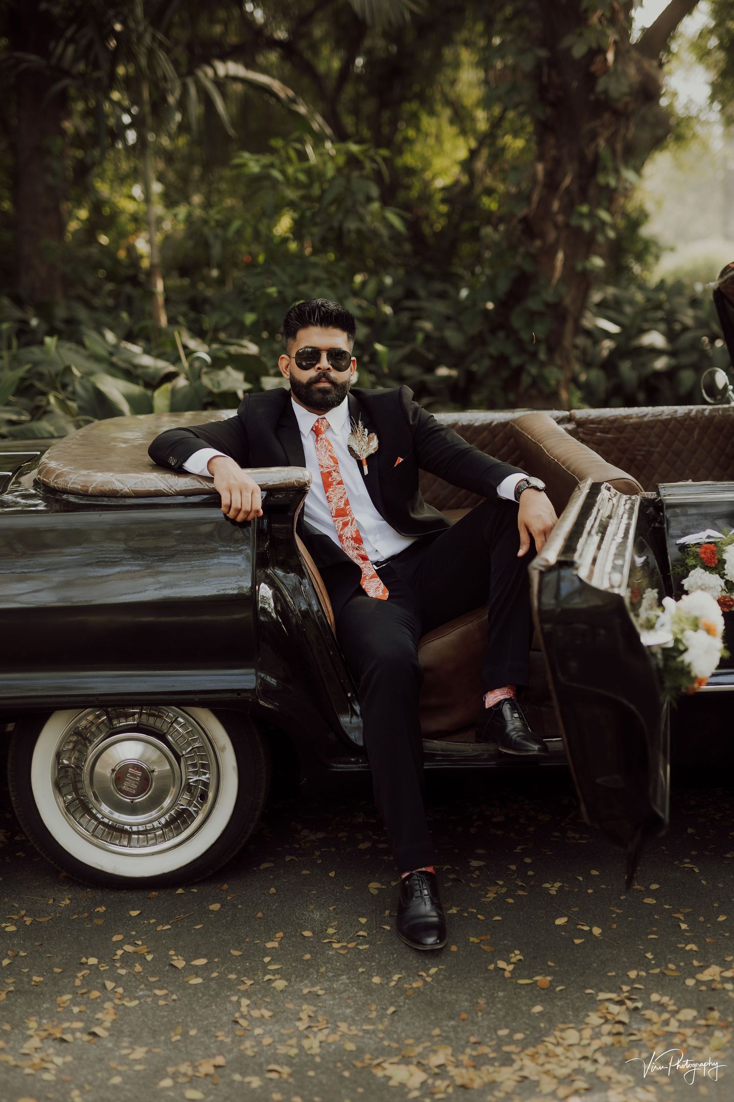 A classic vintage black car with whitewall tires parked in a lush garden setting with someone in a formal suit leaning against it.