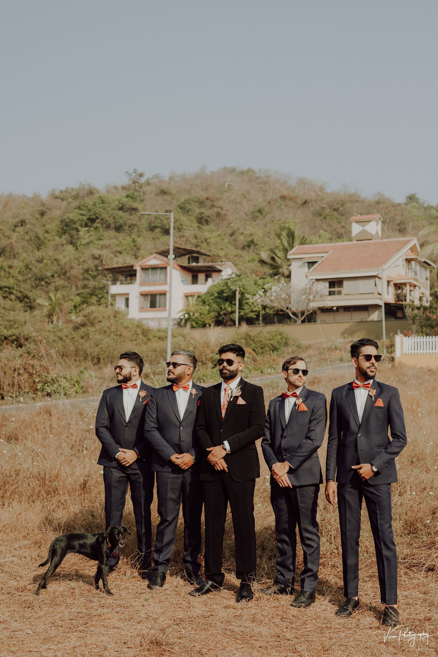 Groomsmen in dark suits stand in a line on a hillside with houses visible in the background.
